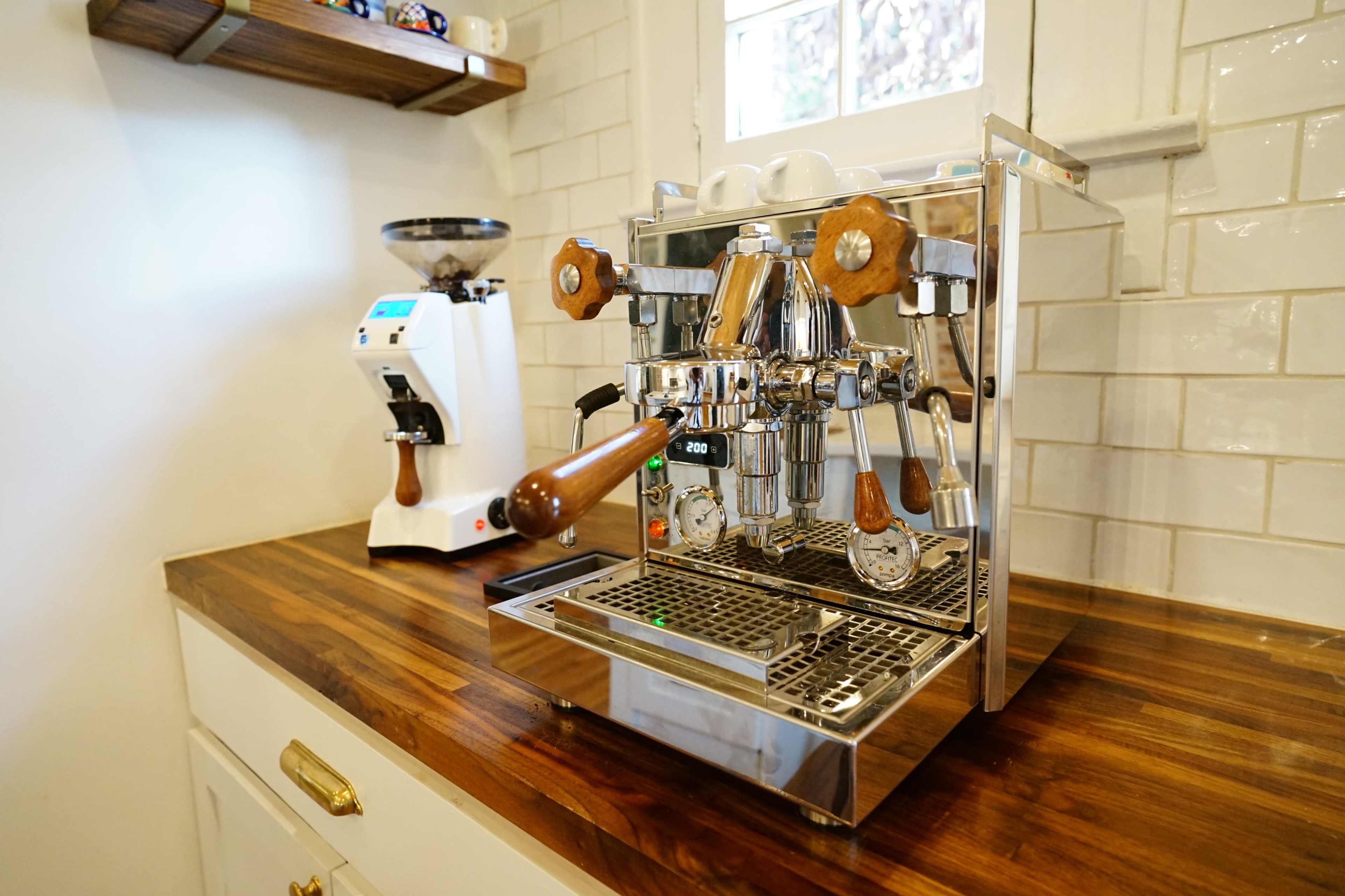 A shiny espresso machine with wooden handles sits on a wooden countertop next to a coffee grinder in a bright kitchen.