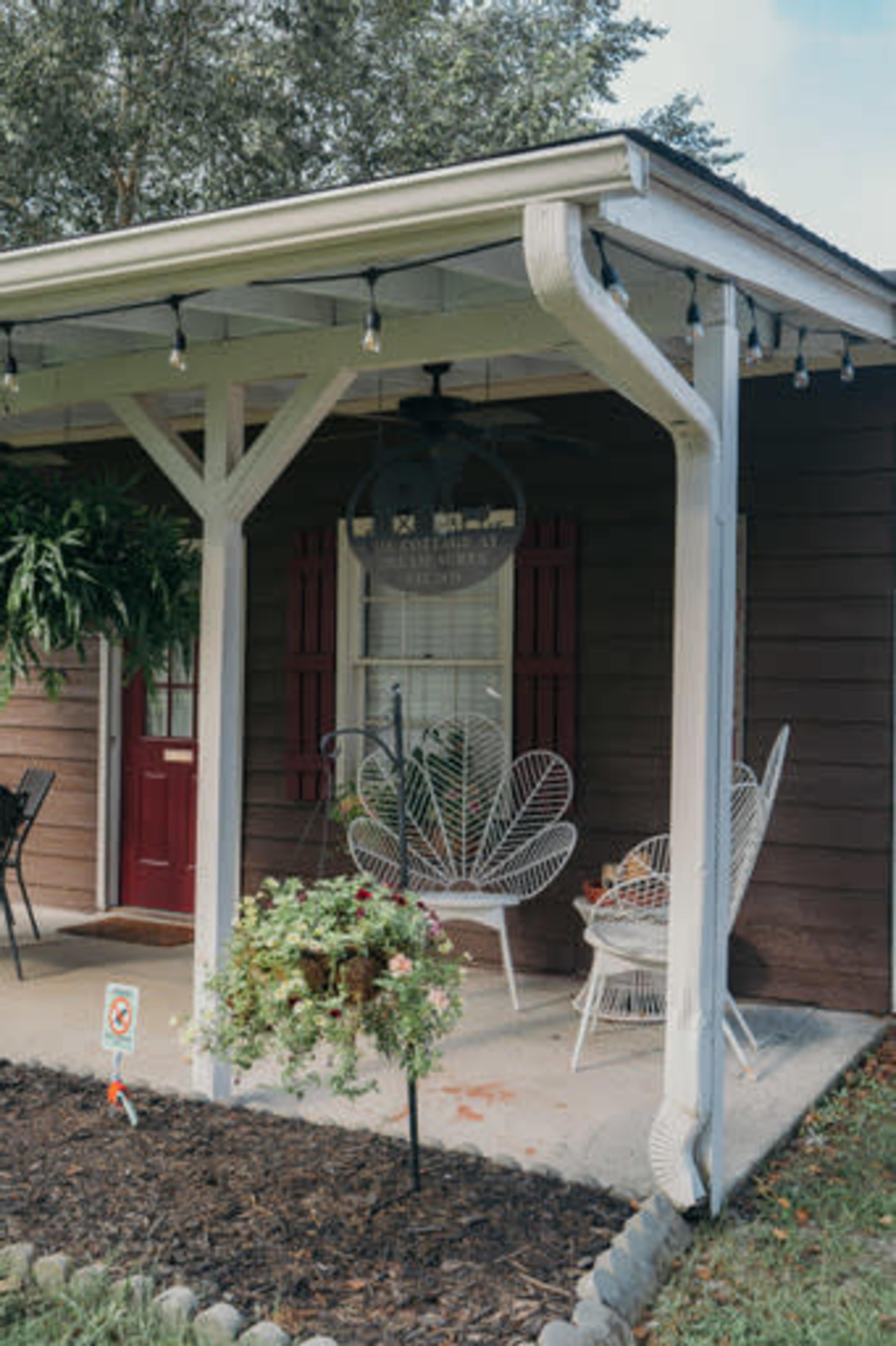 A porch with a ceiling fan features two white wicker chairs and hanging plants, set against a brown wooden house with red shutters.