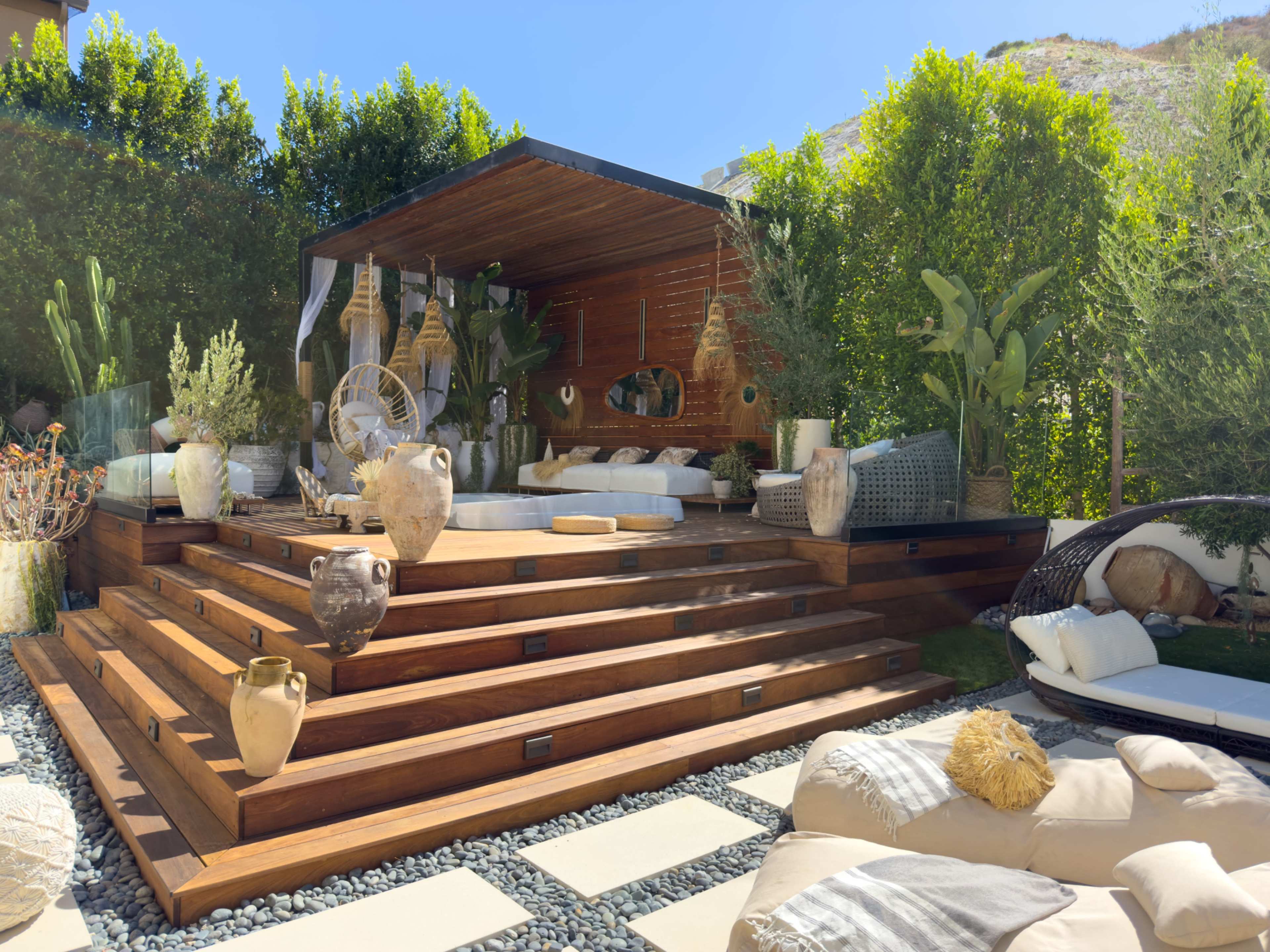 The image shows a modern outdoor living area with wooden steps leading to a covered seating space adorned with plants and decorative items.