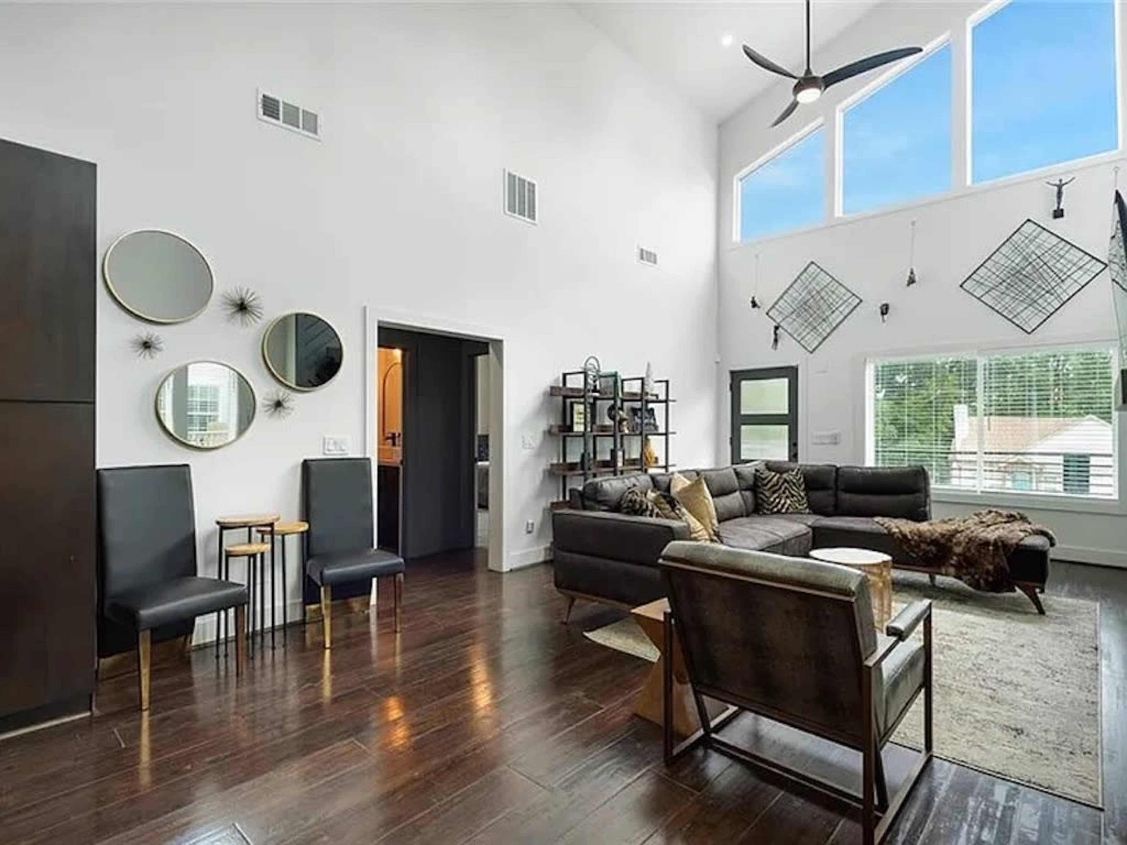 A modern living room features large windows, a sectional sofa, a shelving unit, and decorative mirrors on the walls.