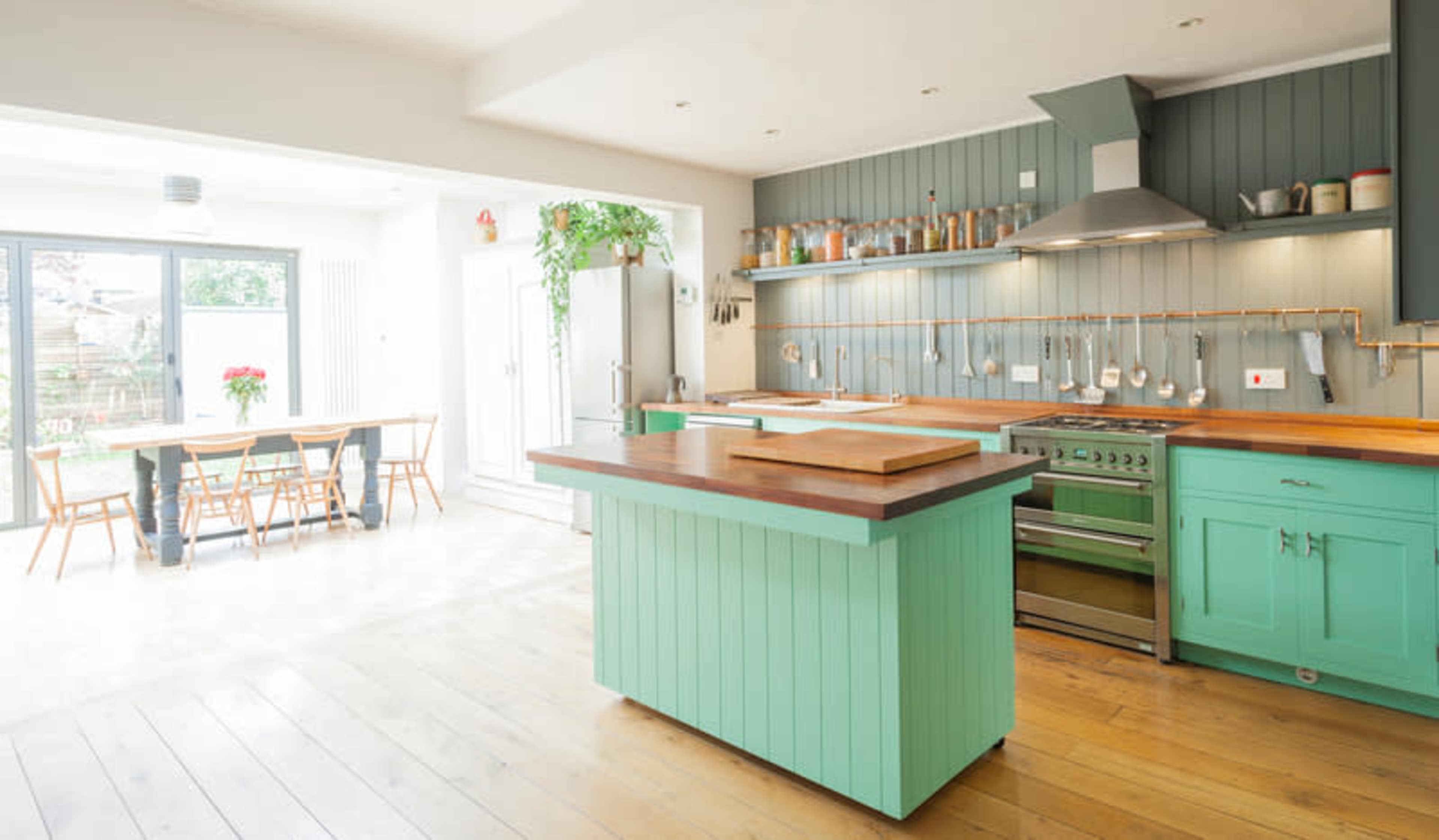 A modern kitchen features a mint green island with a wooden countertop, stainless steel appliances, and a dining area illuminated by natural light from large windows.