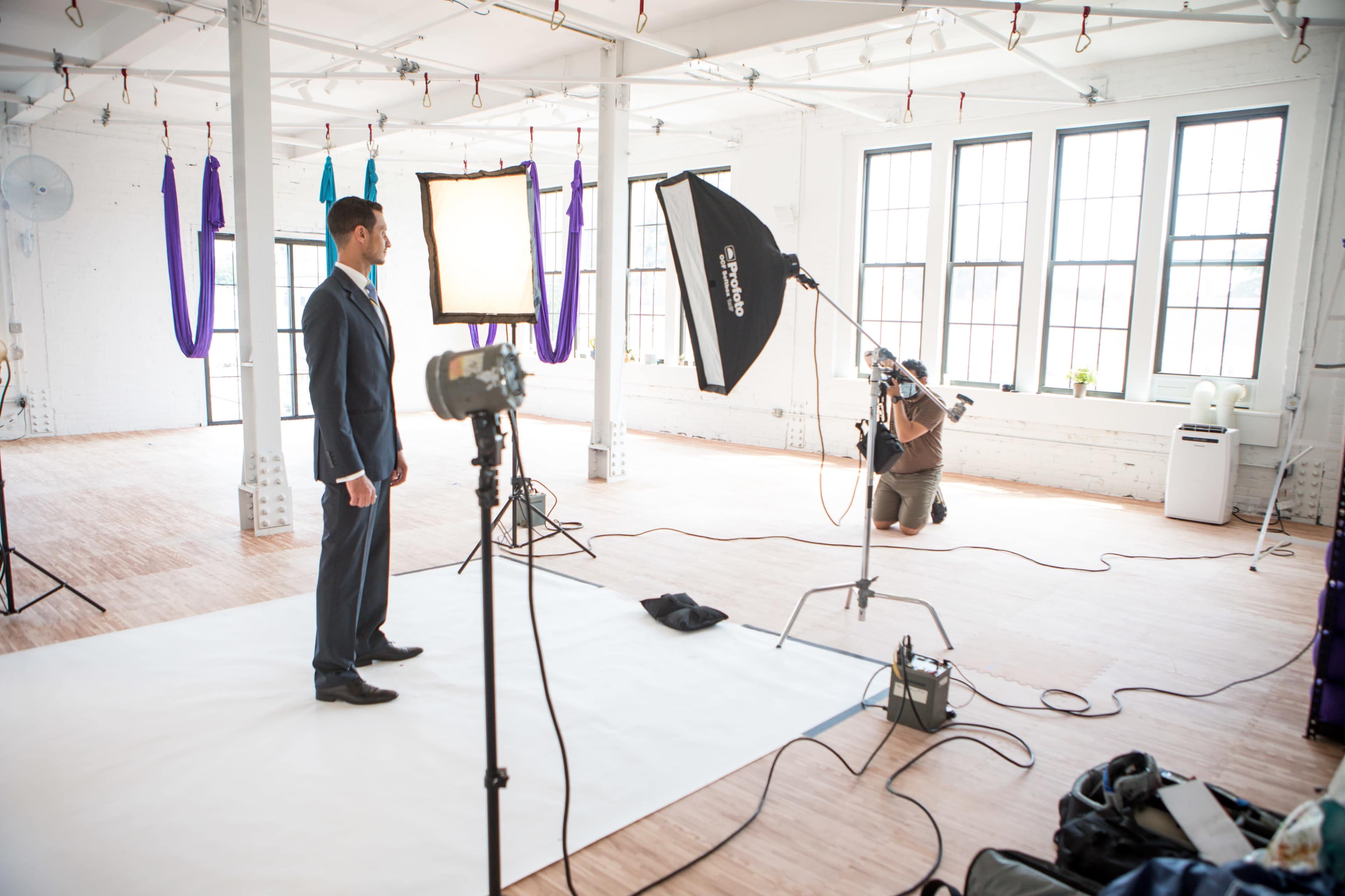 A man in a suit stands on a white backdrop, while a photographer kneels behind a camera and softbox in a well-lit studio.