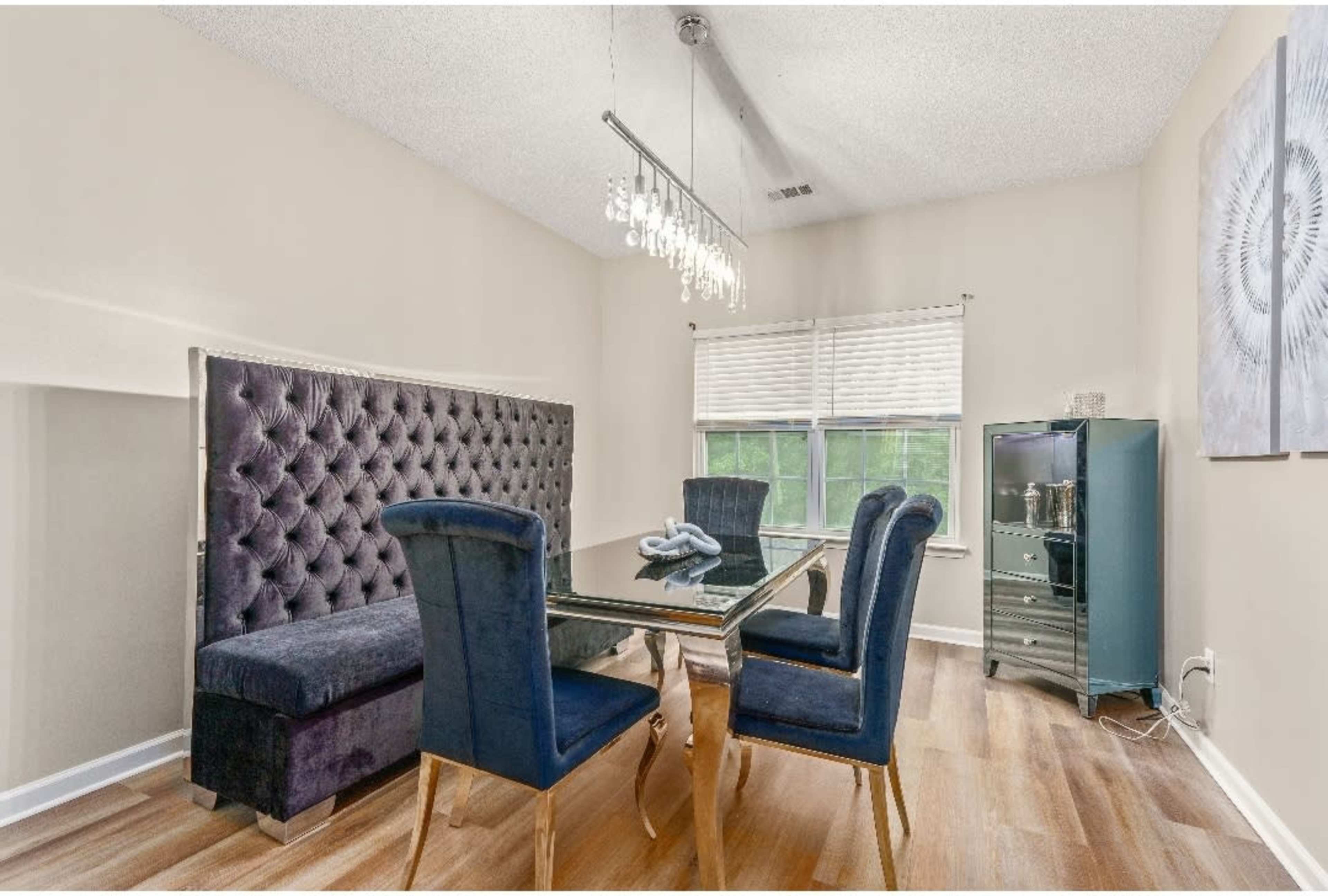A dining area features a tufted booth and four blue chairs around a glass table, illuminated by a modern chandelier.