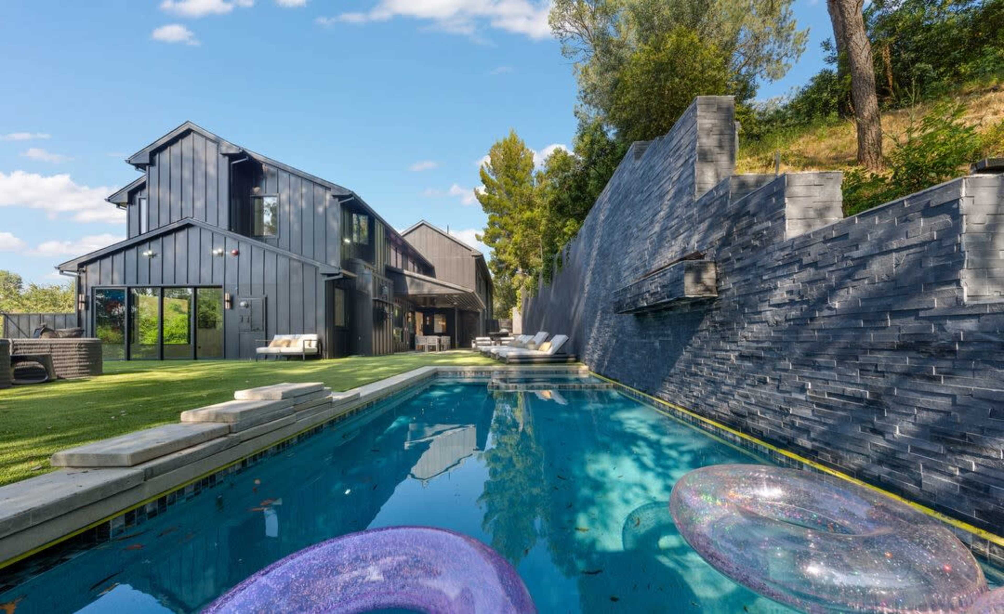 A modern house with a black exterior overlooks a swimming pool, which features inflatable rings, surrounded by a stone wall and greenery.