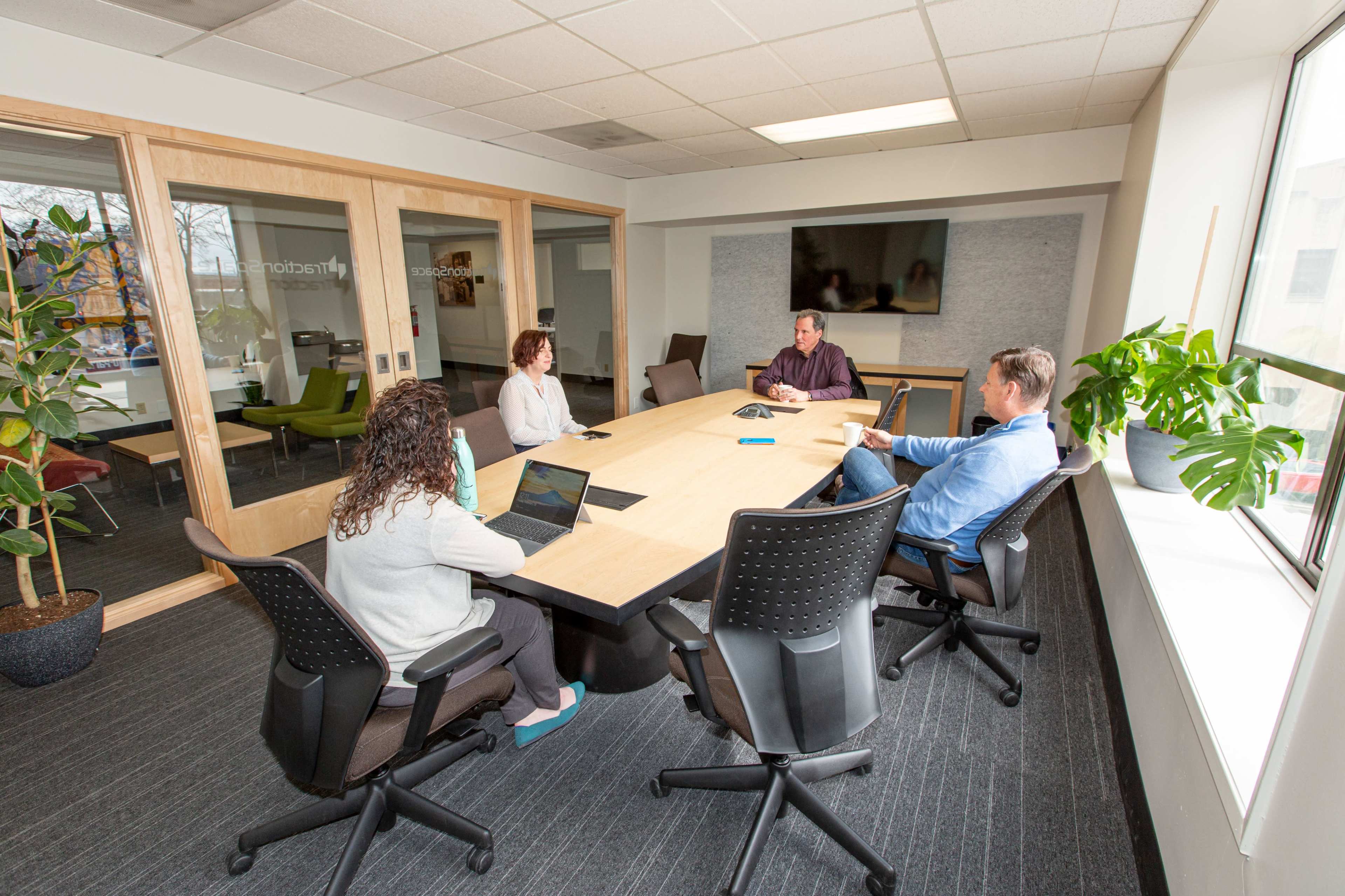 A group of four people are seated around a rectangular conference table in a modern meeting room, with laptops and documents in front of them.
