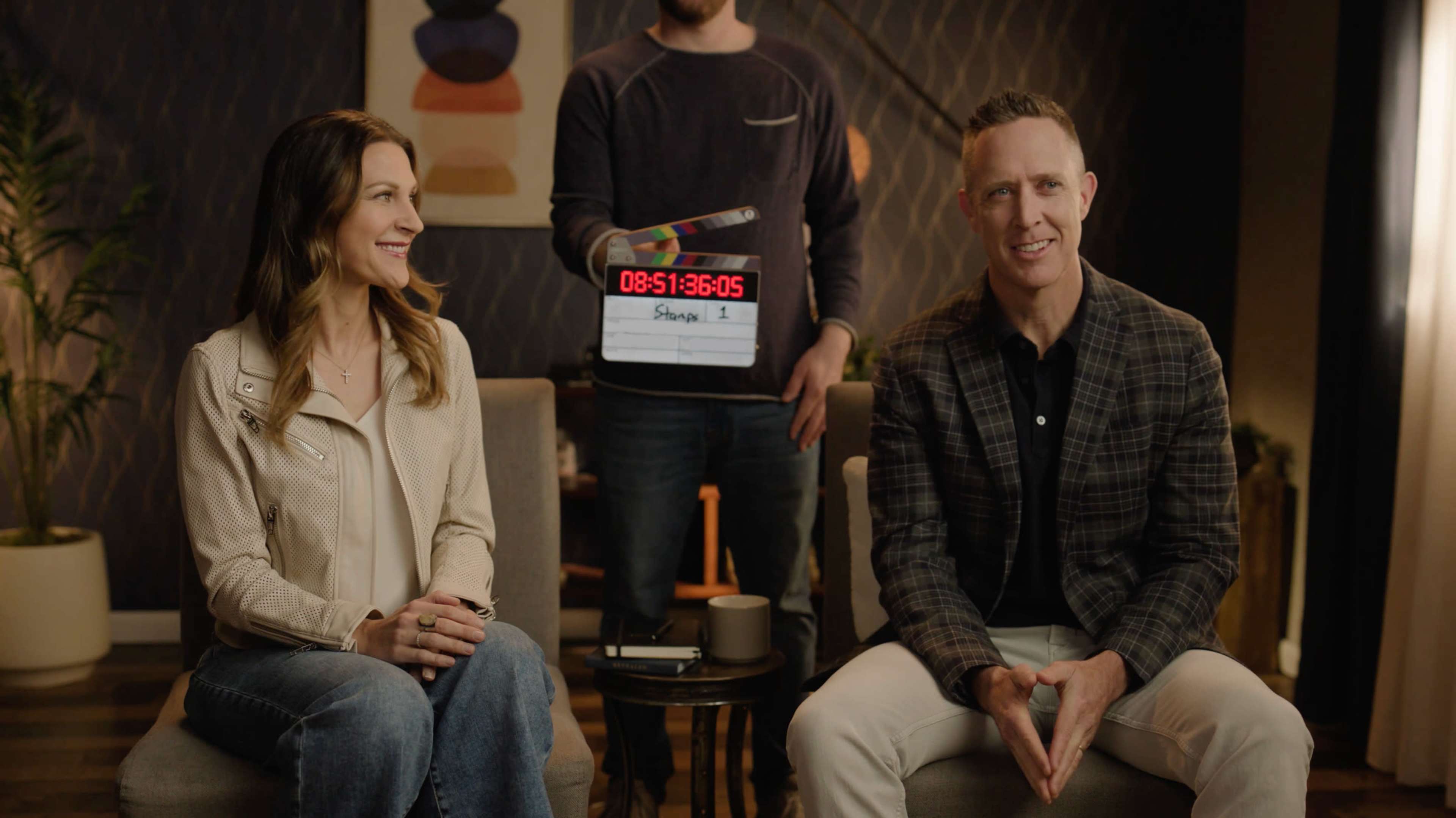 Two seated individuals are smiling at the camera while a person holds a film slate behind them, indicating the start of a recording session.