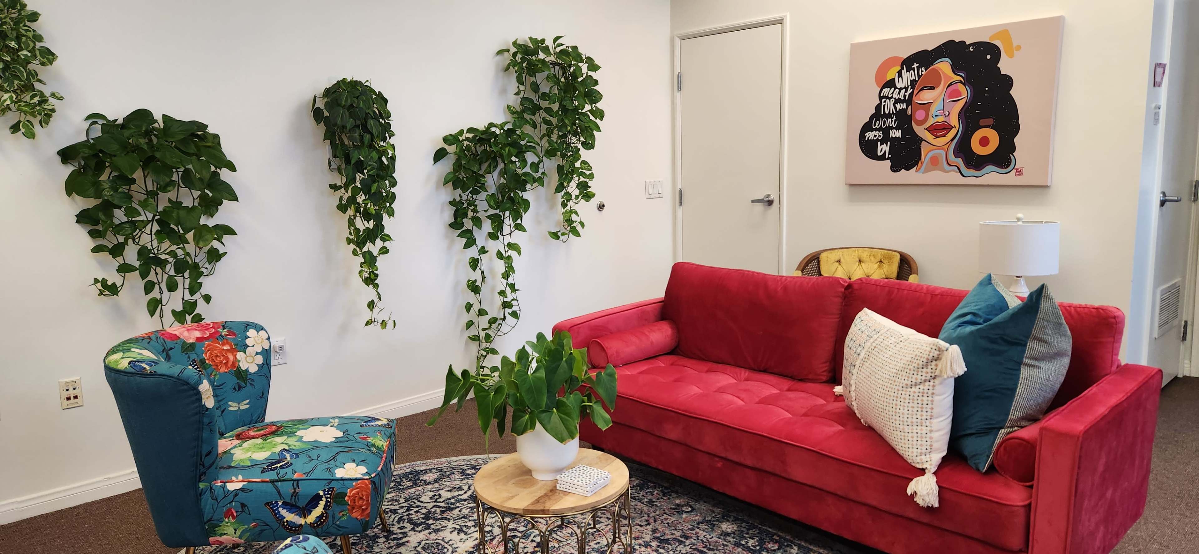 A bright living room with a red couch, a colorful chair, a small wooden table with a plant, and two wall-mounted greenery pieces, accompanied by a vibrant artwork on the wall.