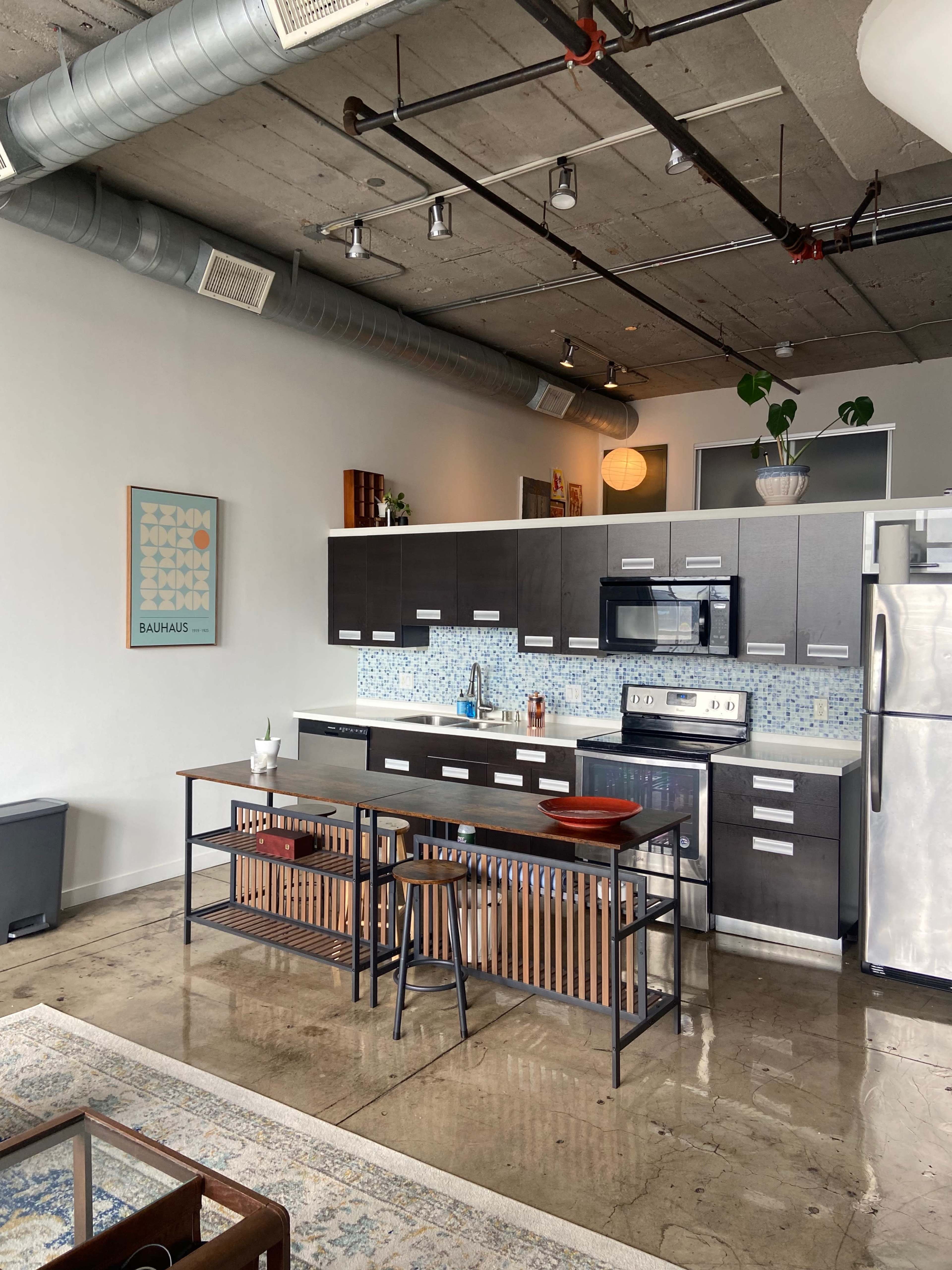 The image shows a modern kitchen with dark cabinetry, a stainless steel stove, and a wooden dining table, set against a concrete wall.