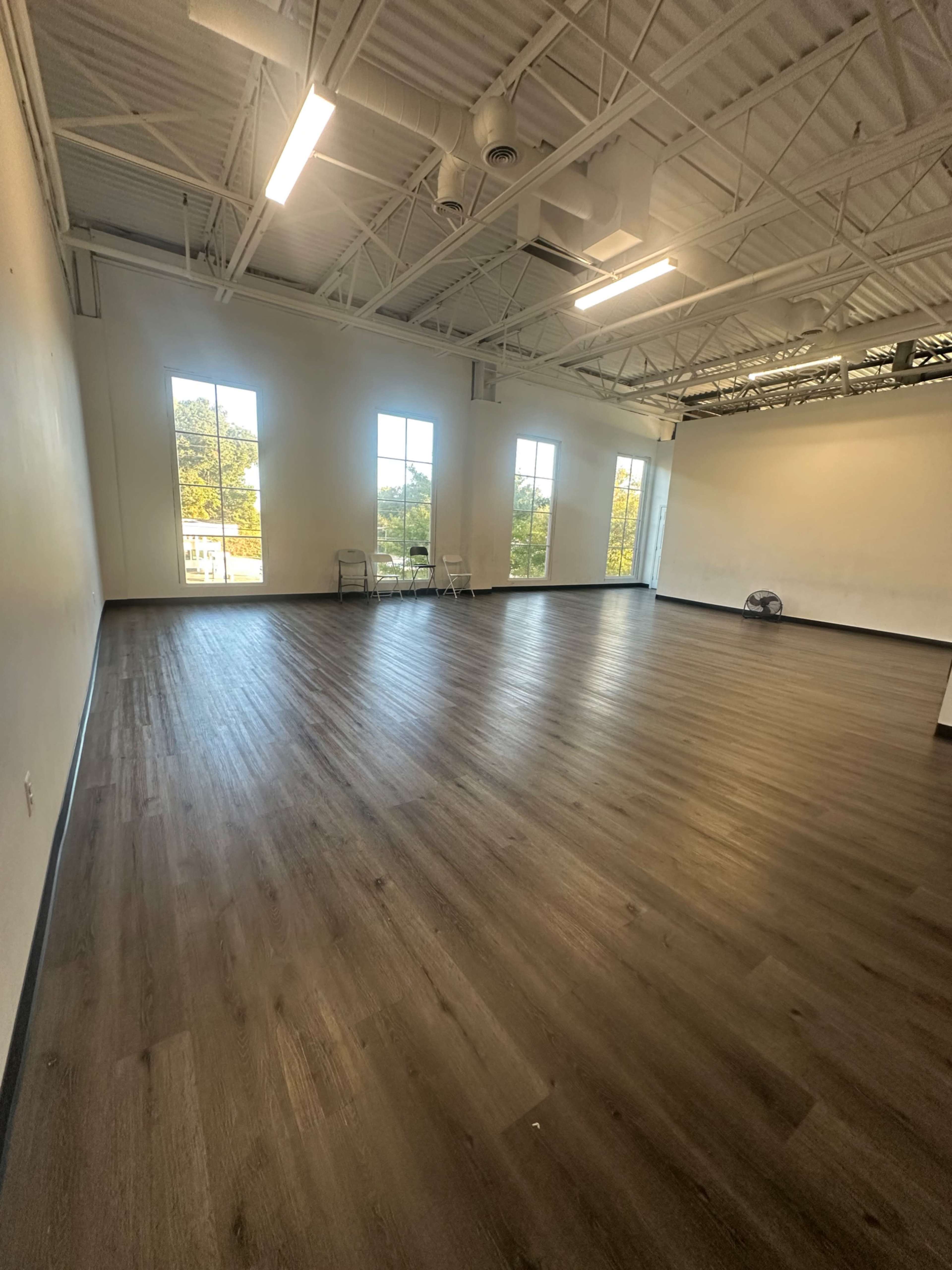 The image shows a spacious room with wooden flooring, large windows allowing natural light, and minimal furniture.
