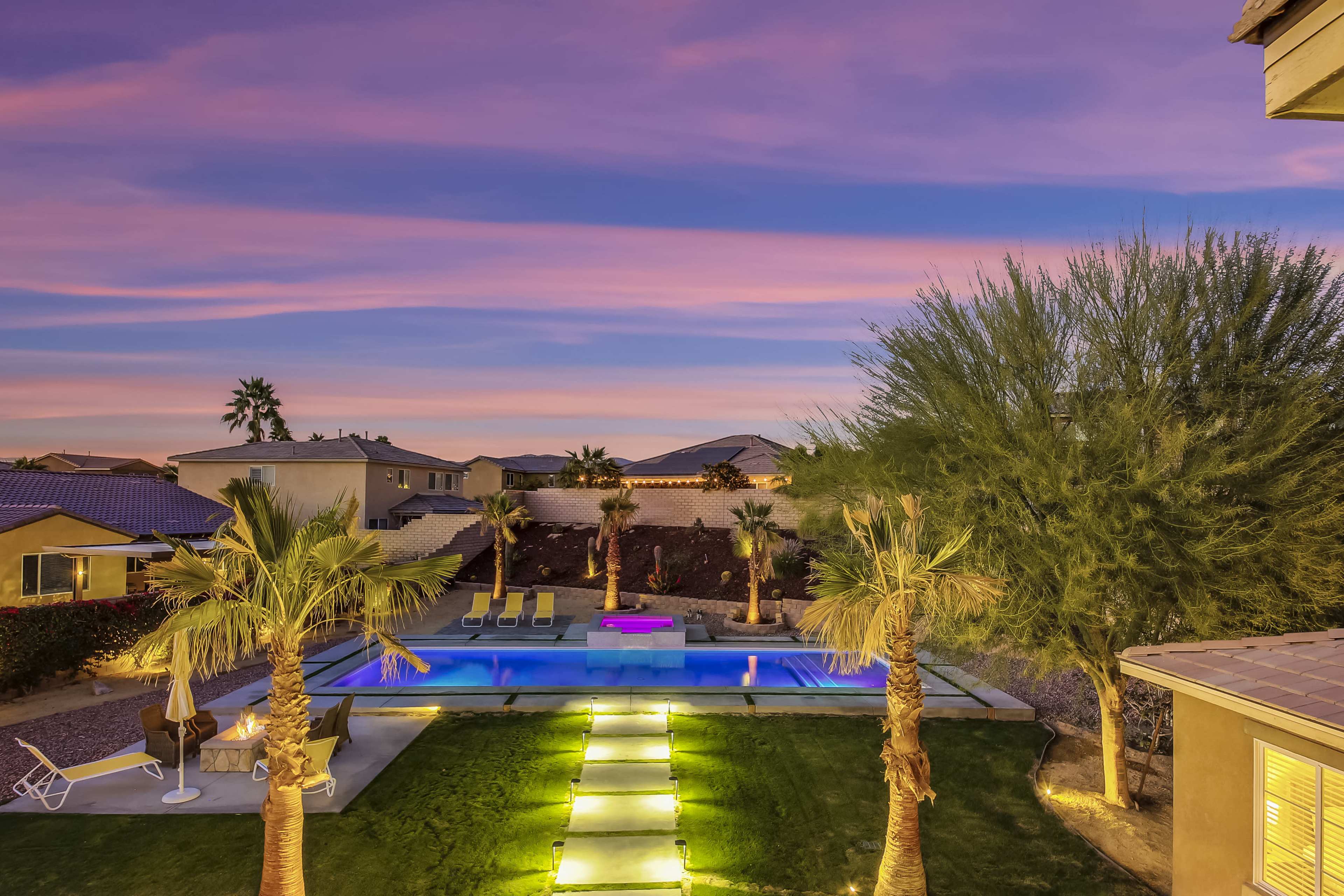 A backyard pool area is illuminated at dusk with palm trees and a colorful sky in the background.