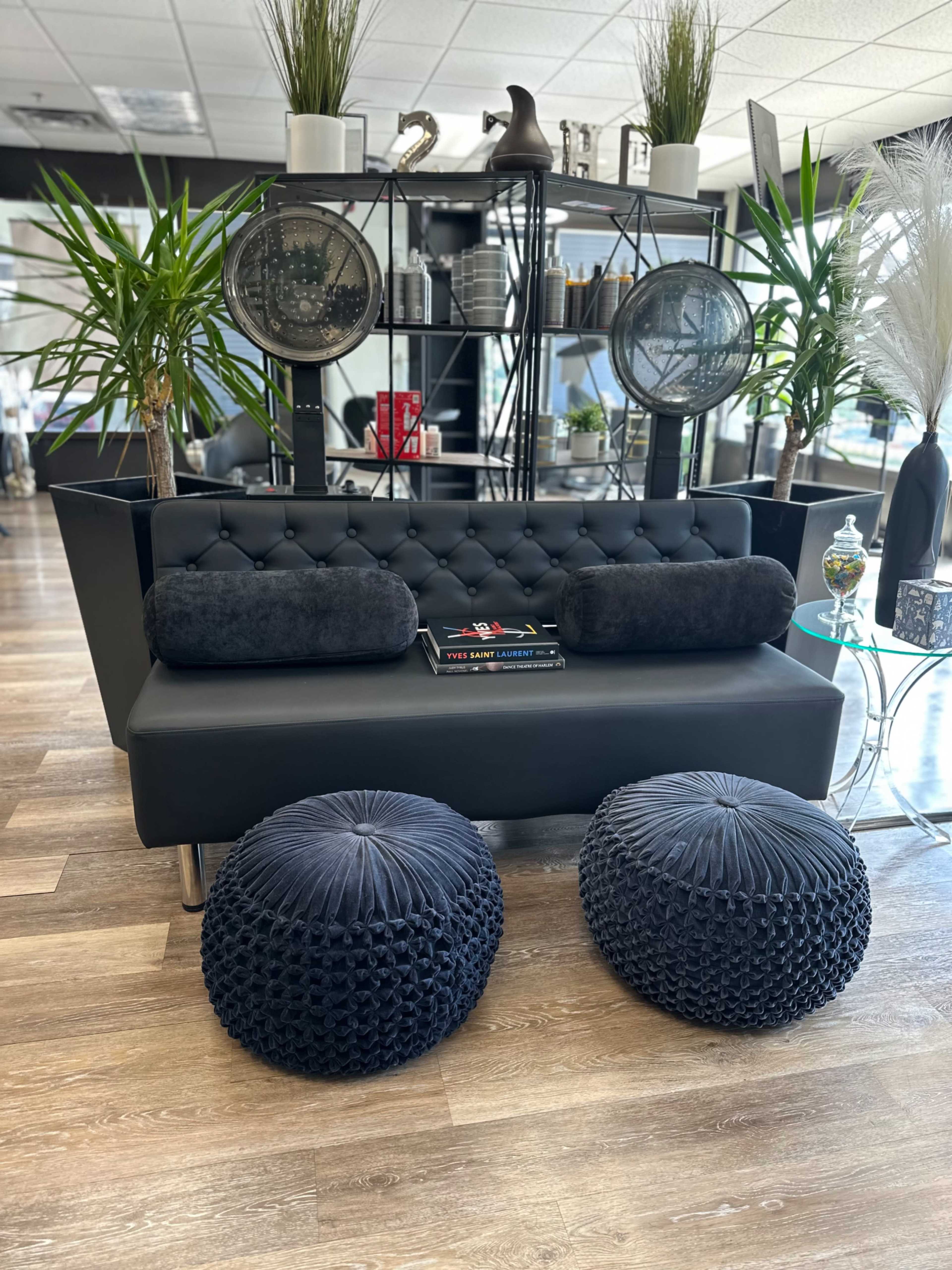 A modern black sofa sits in a stylish interior, accompanied by round black poufs and decorative plants, with shelves displaying various items in the background.