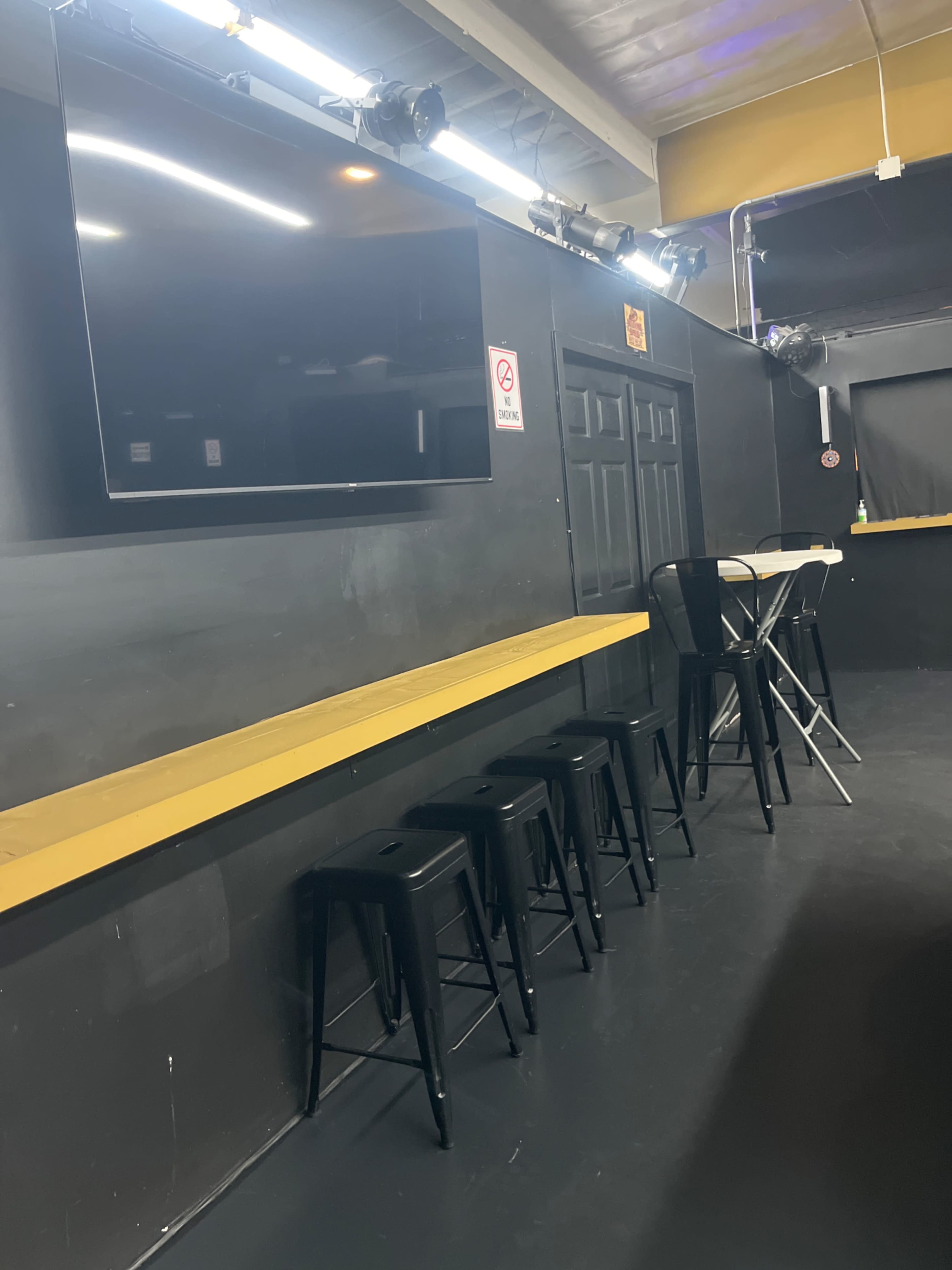 The image shows a dimly lit venue with black walls, a long yellow bar counter, high black stools, and a television mounted on the wall.