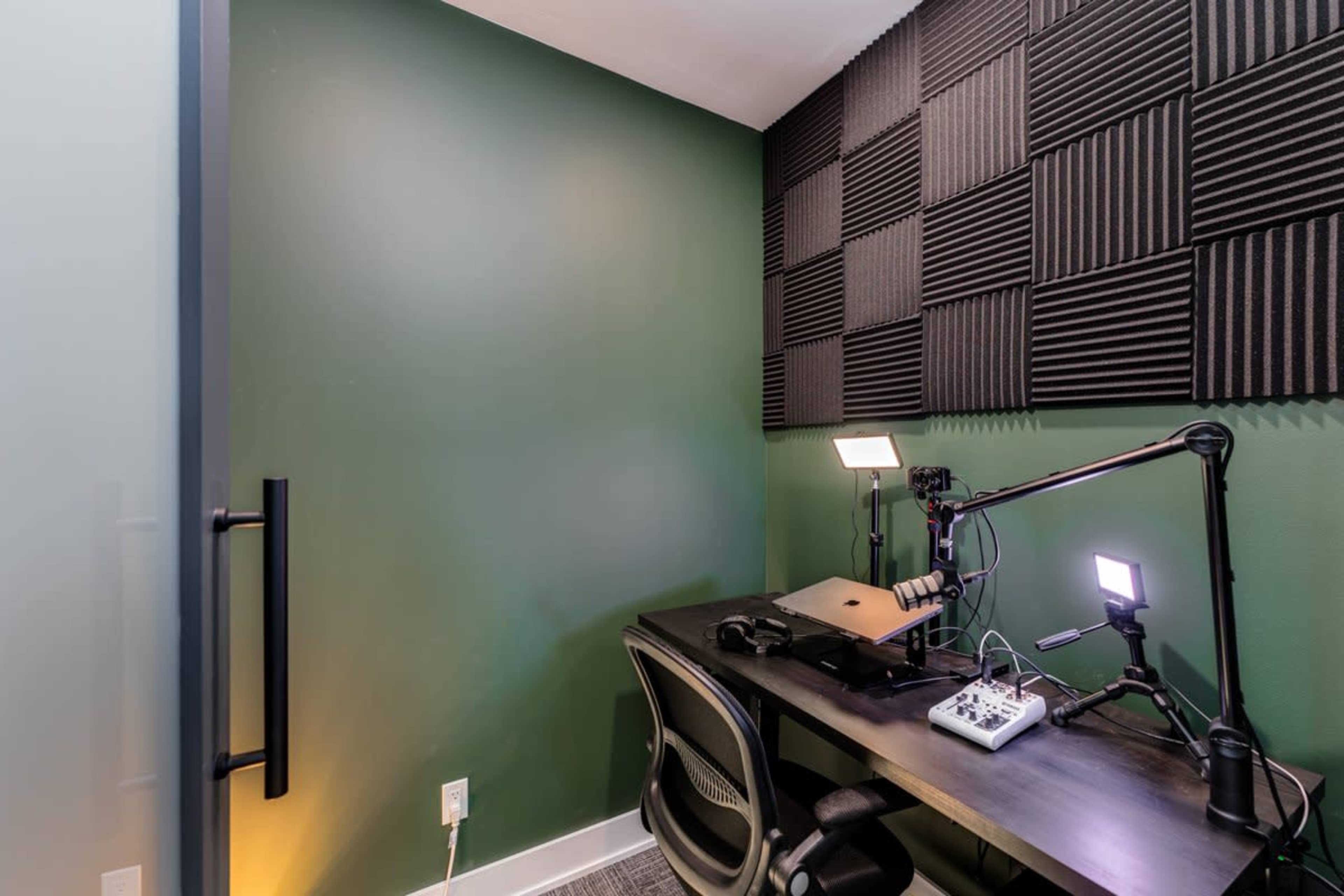 The image shows a small recording studio featuring a dark green wall, acoustic foam panels, and a desk with microphones and lighting equipment.