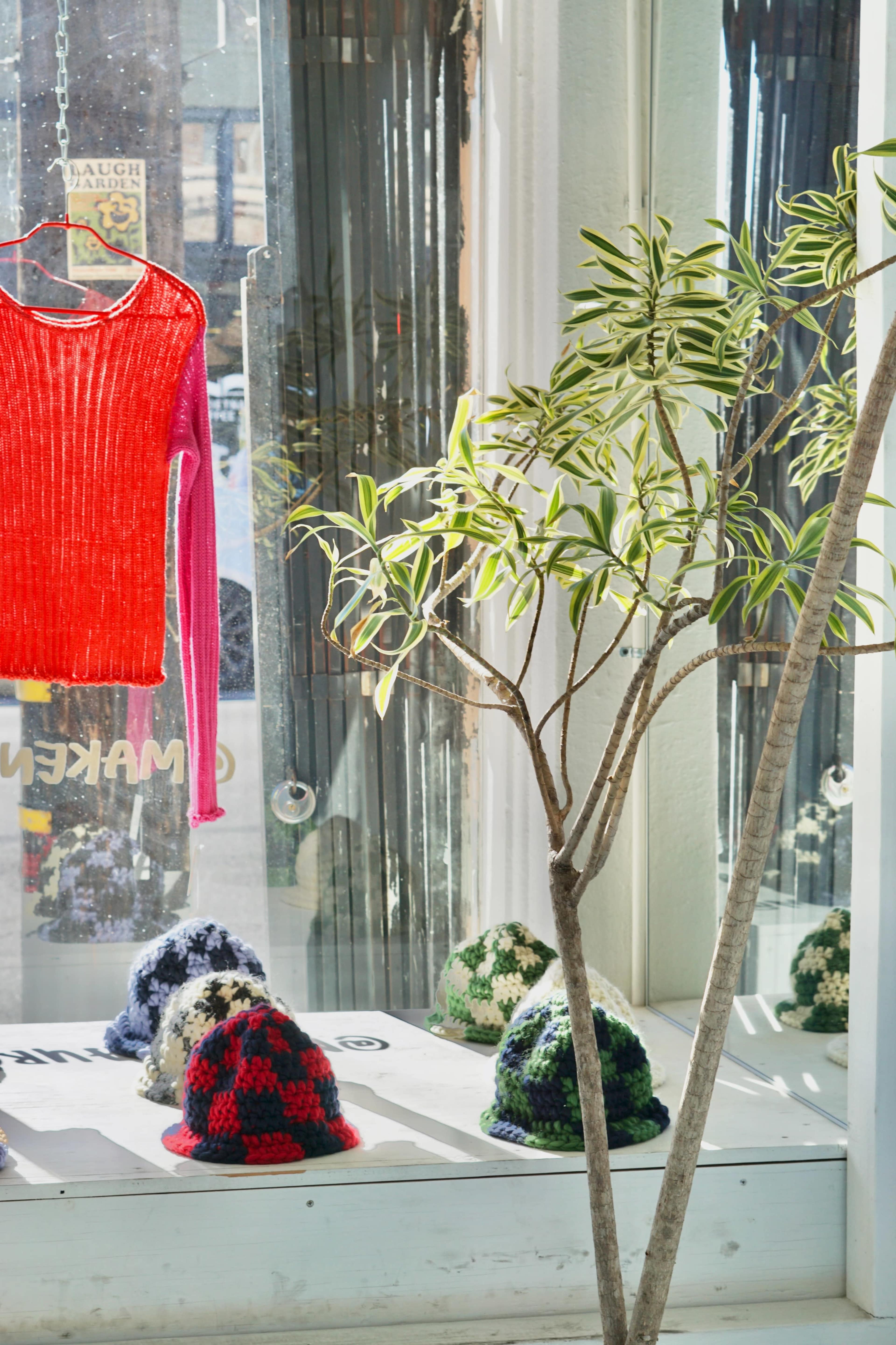 A red and pink sweater hangs in a window display beside a potted plant and several colorful knitted hats on a white surface.