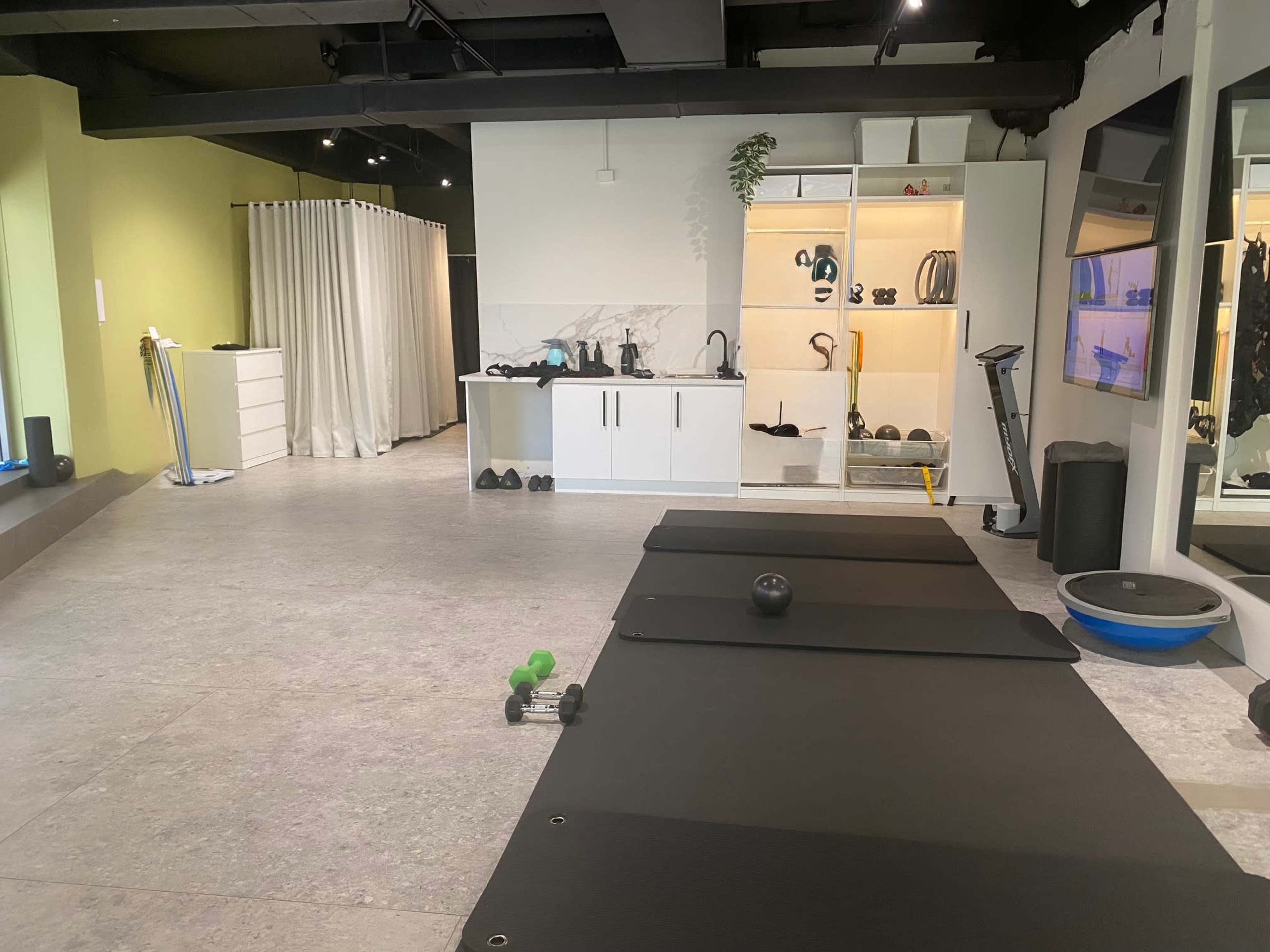 The image shows a modern fitness studio with exercise mats, weights, and a kitchenette area featuring shelves and various fitness equipment.