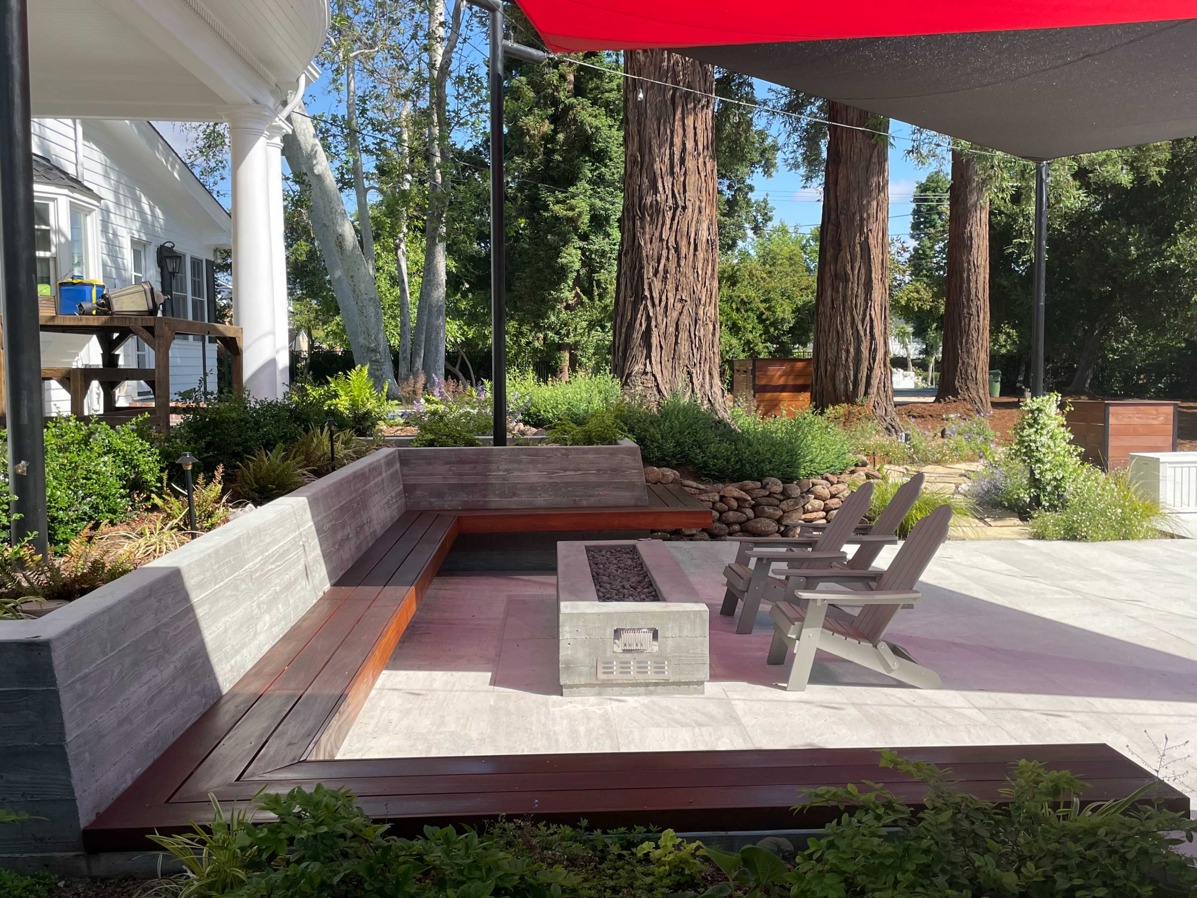 A modern outdoor seating area with a fire pit and two wooden chairs, surrounded by greenery and tall trees.