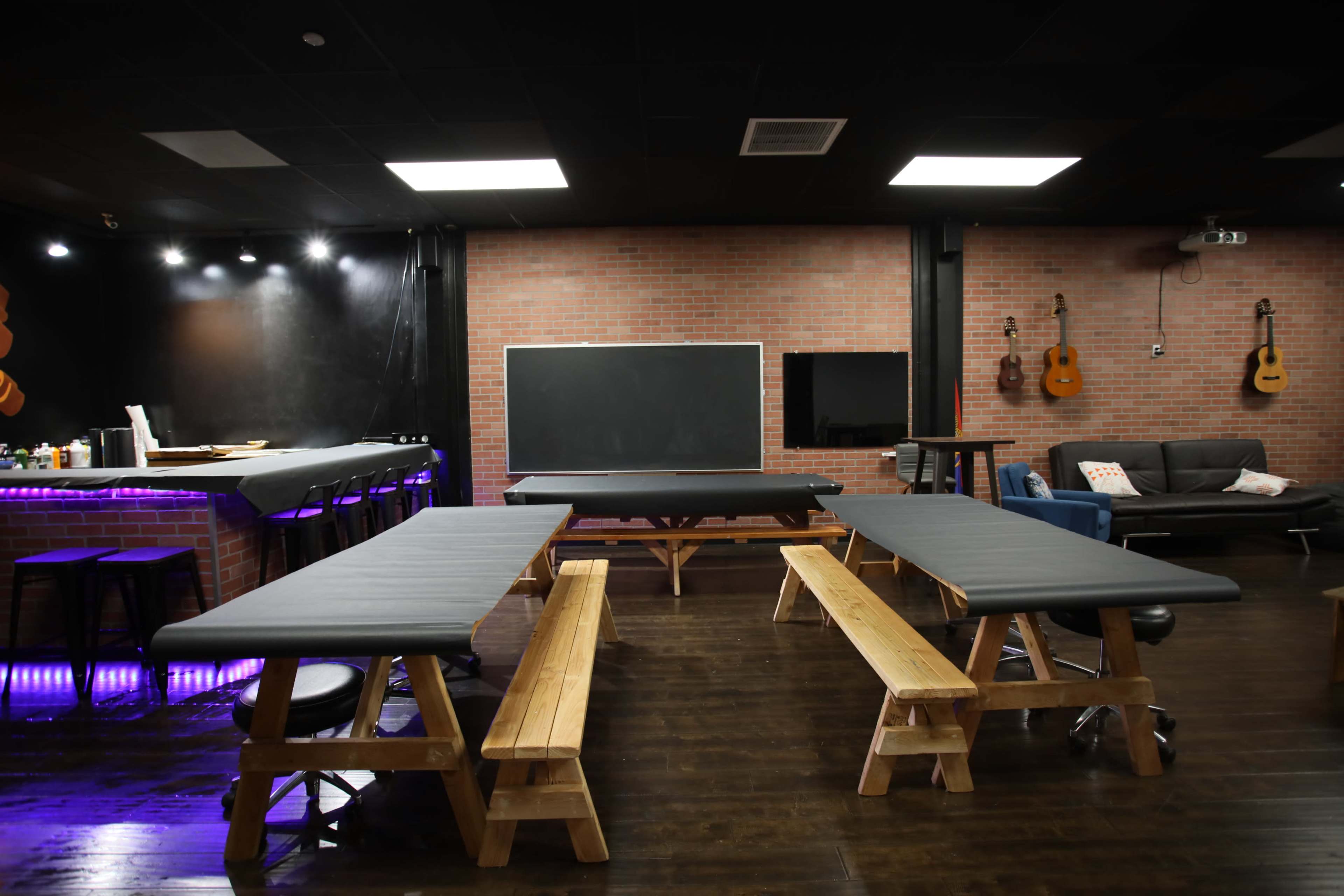 The image shows a room with two large wooden tables surrounded by benches, a bar area with purple lighting, and a chalkboard on one wall.