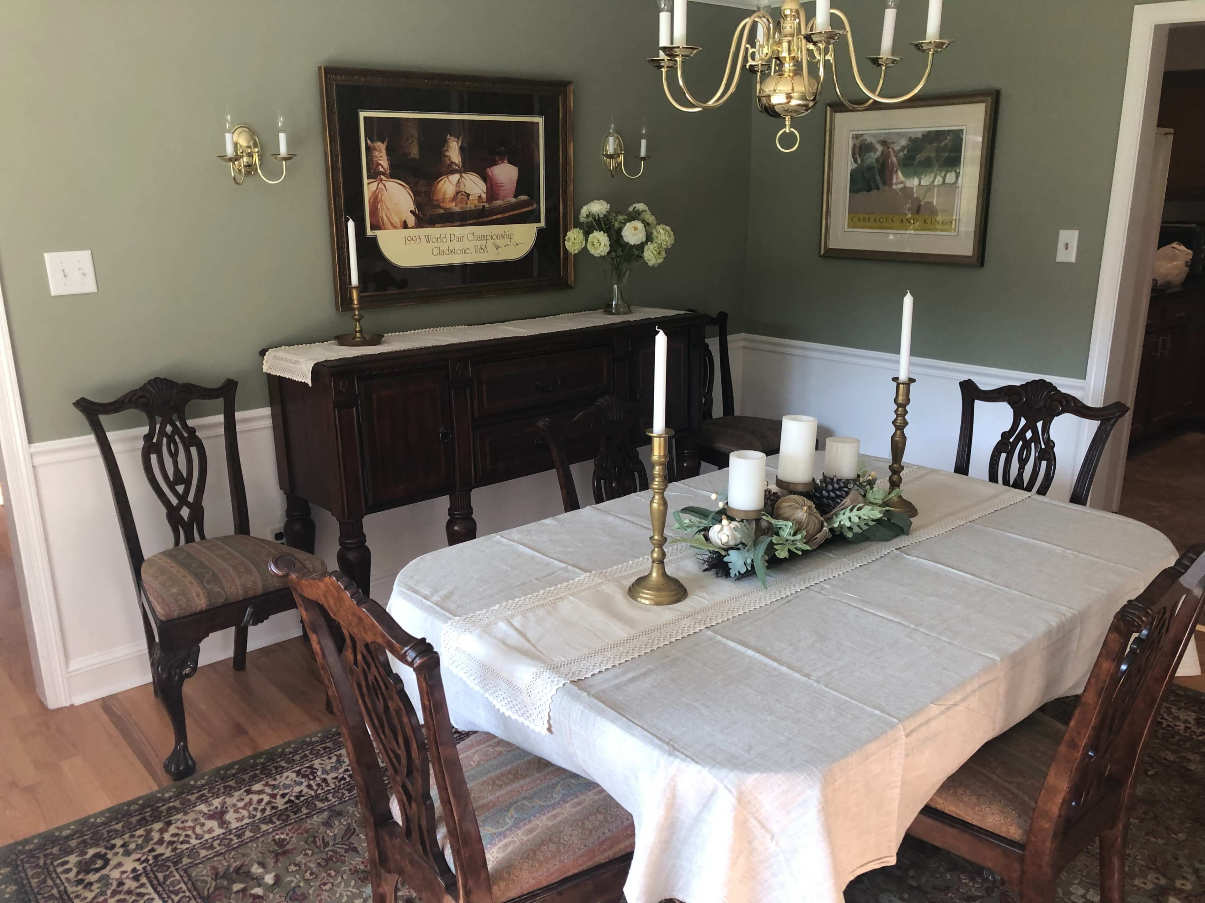 A dining room features a wooden table with a cream-colored tablecloth, surrounded by chairs, and a sideboard adorned with decorative items and artwork on the walls.