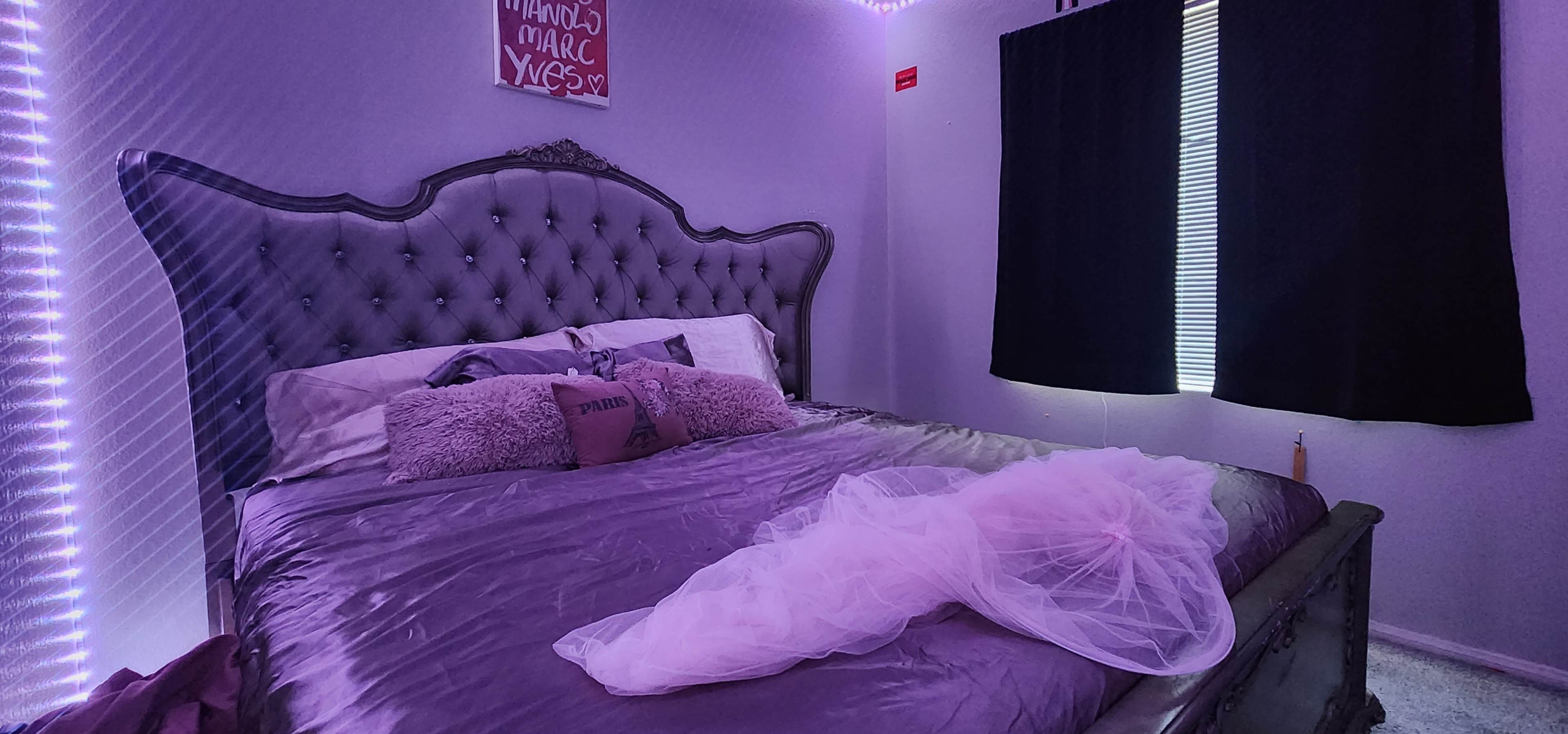 A bedroom with a tufted gray headboard, a purple bedspread, and sheer fabric draped over the bed, illuminated by soft lighting along the walls.