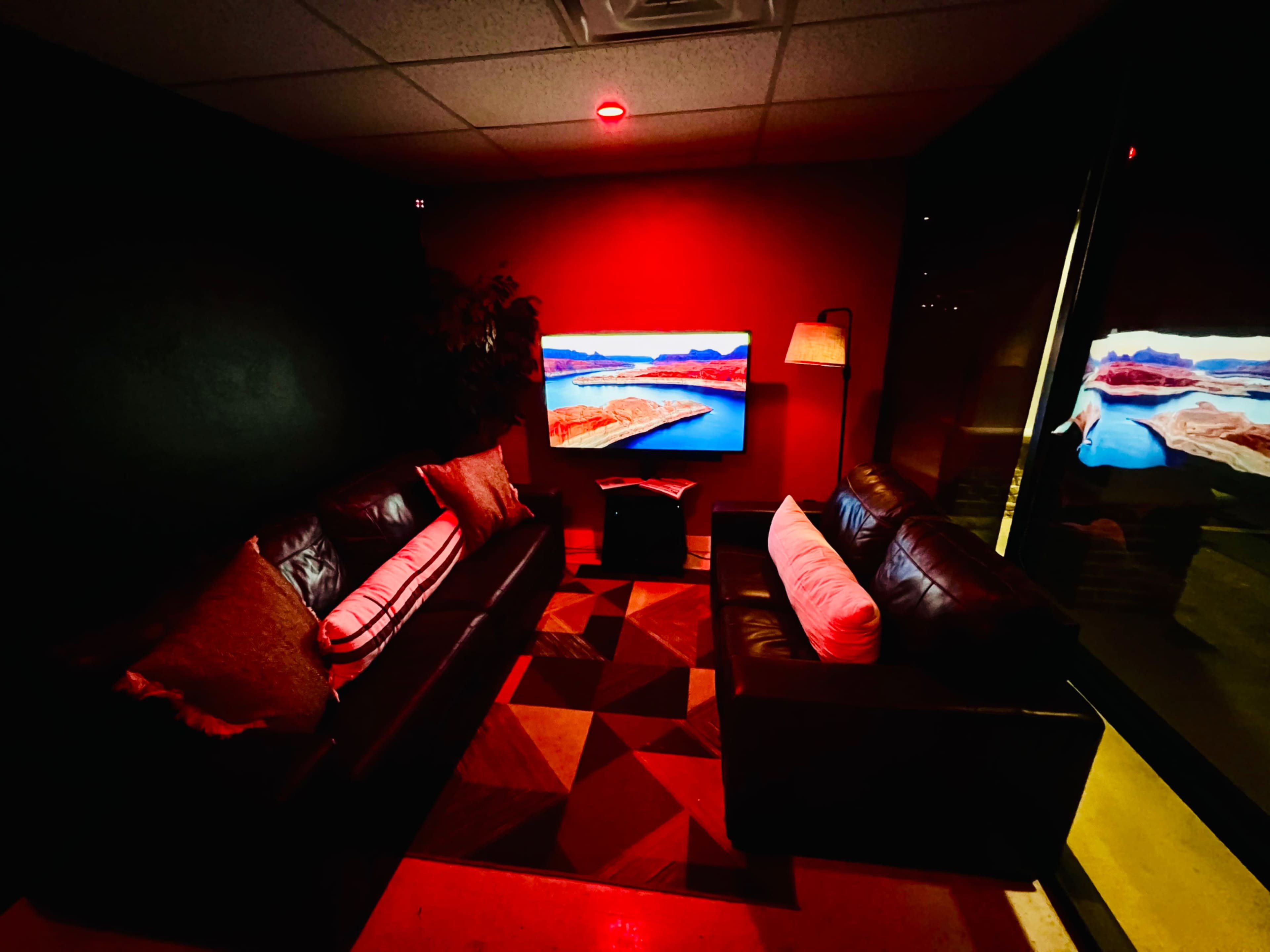 A small lounge area features two black leather sofas, a coffee table, and a television displaying a landscape, all illuminated by red lighting.