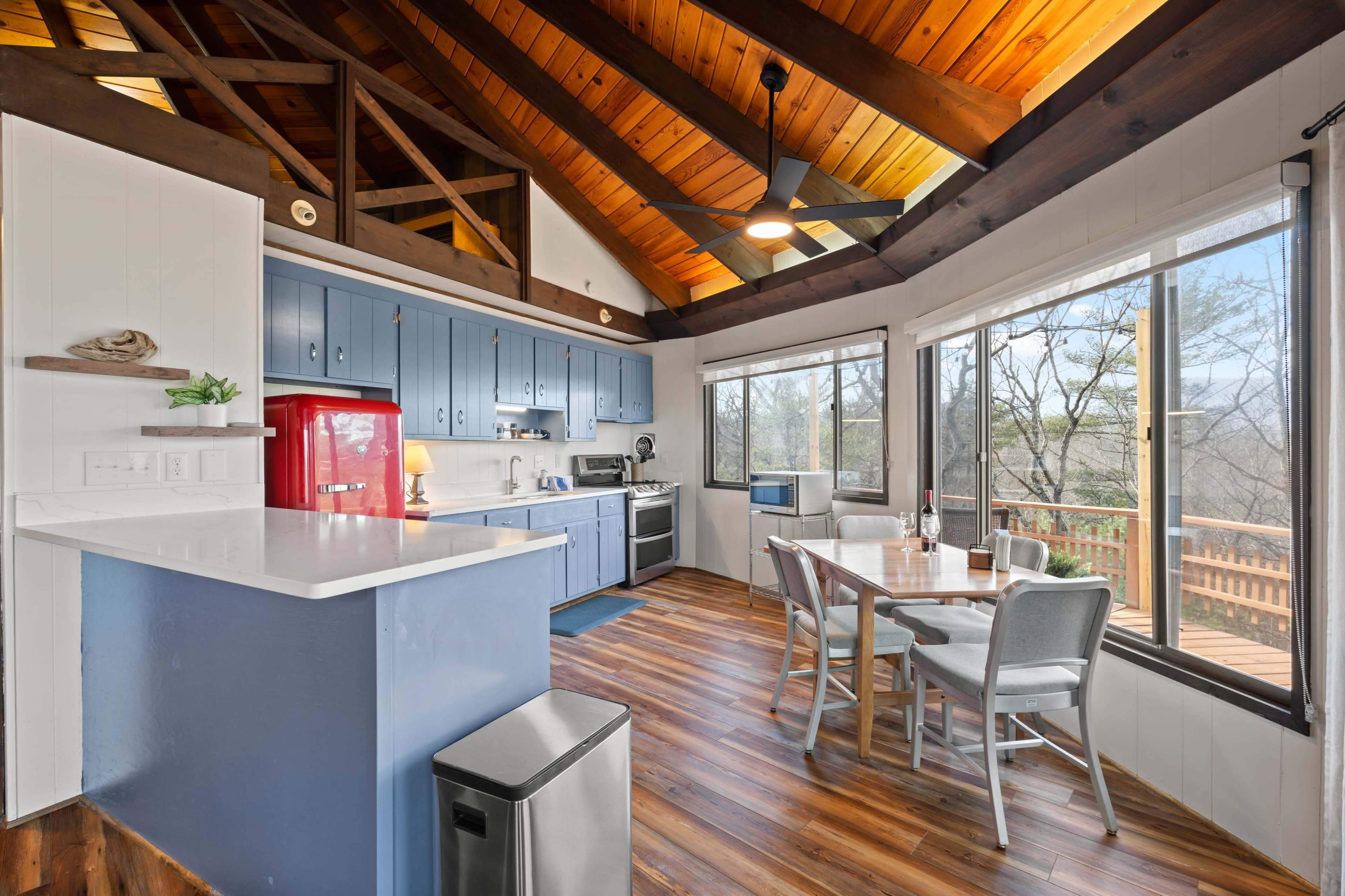 The image shows a modern kitchen with blue cabinetry, a red refrigerator, and a dining area featuring a round table and large windows.