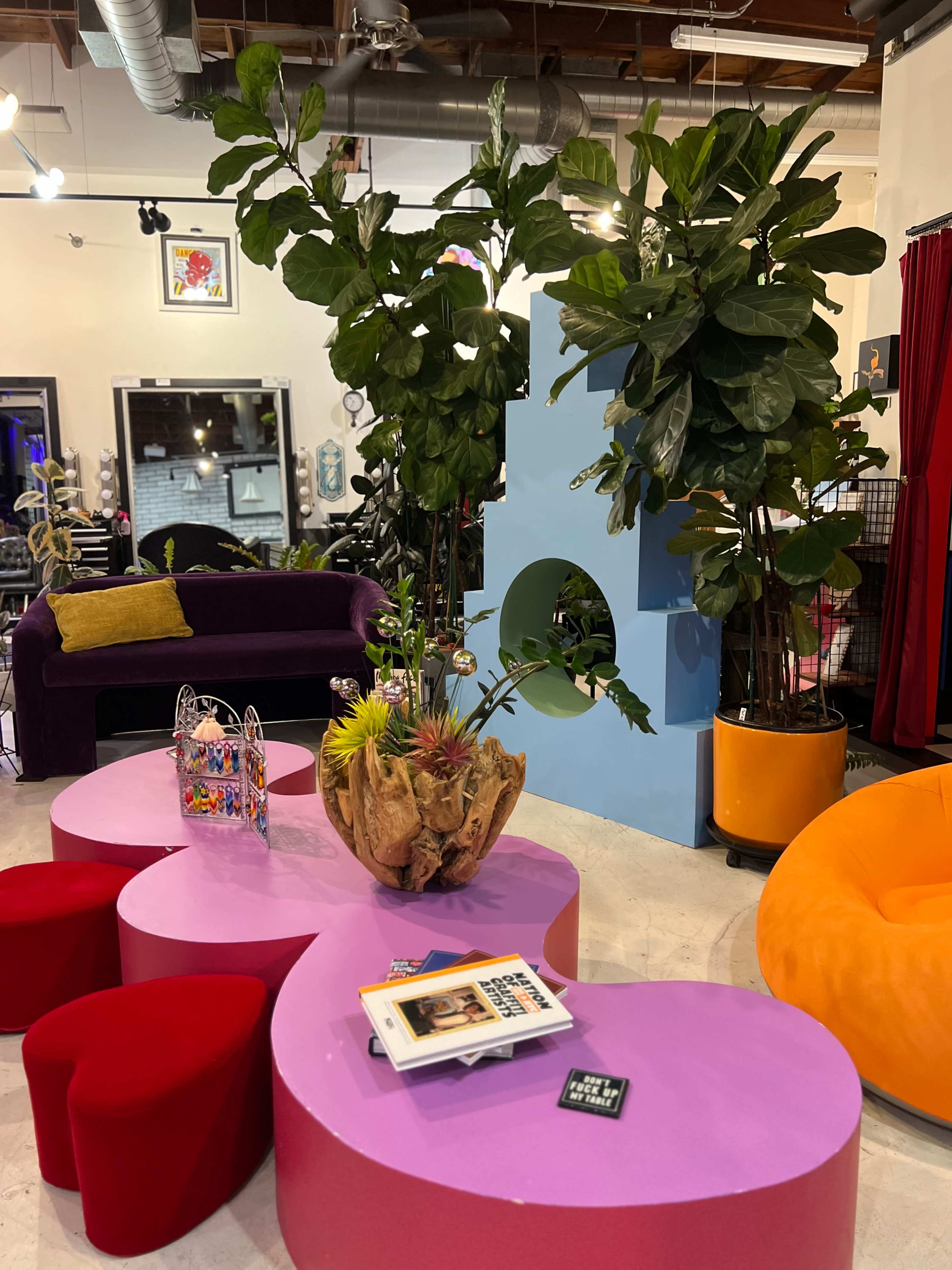 A colorful lounge area features a pink table, red heart-shaped stools, a wooden planter with succulents, tall green plants, and various seating options.