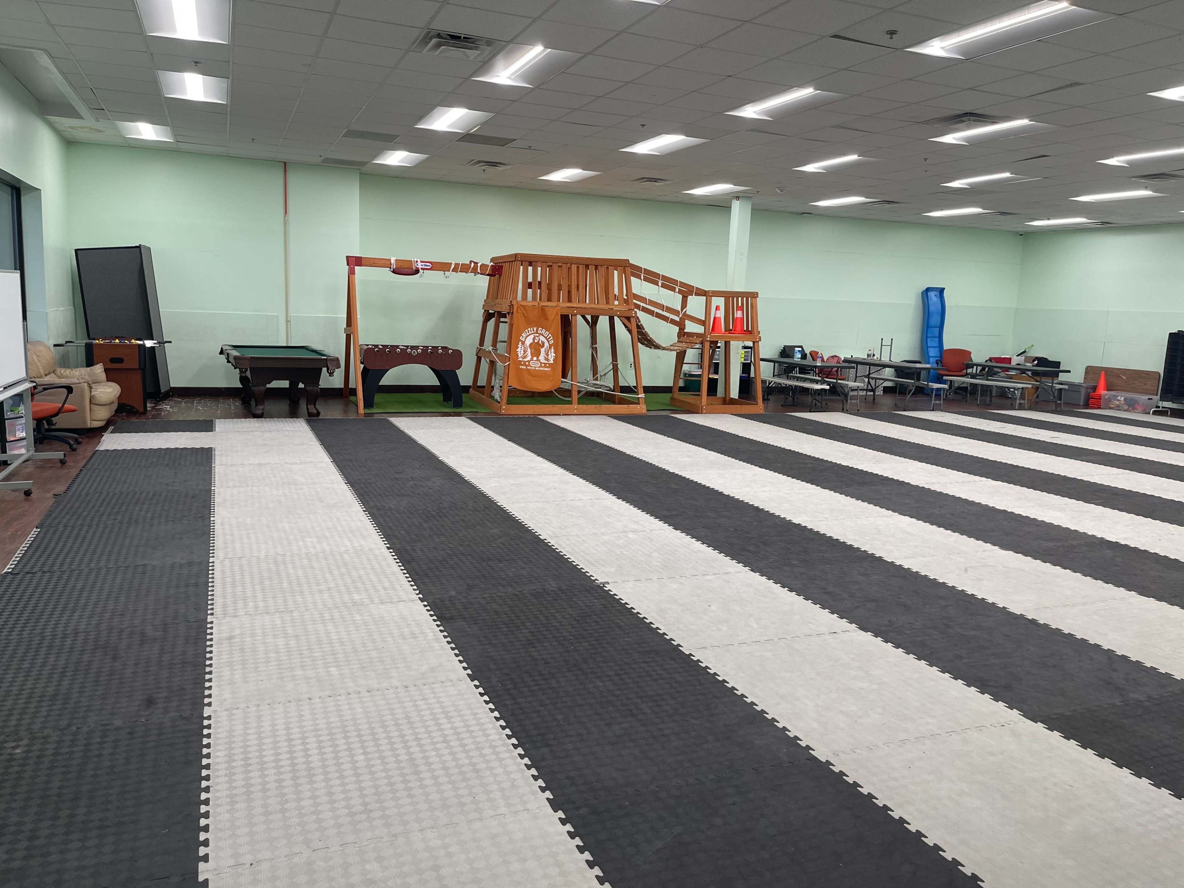 A large room with alternating black and white mats, featuring a playground structure and various furniture along the walls.