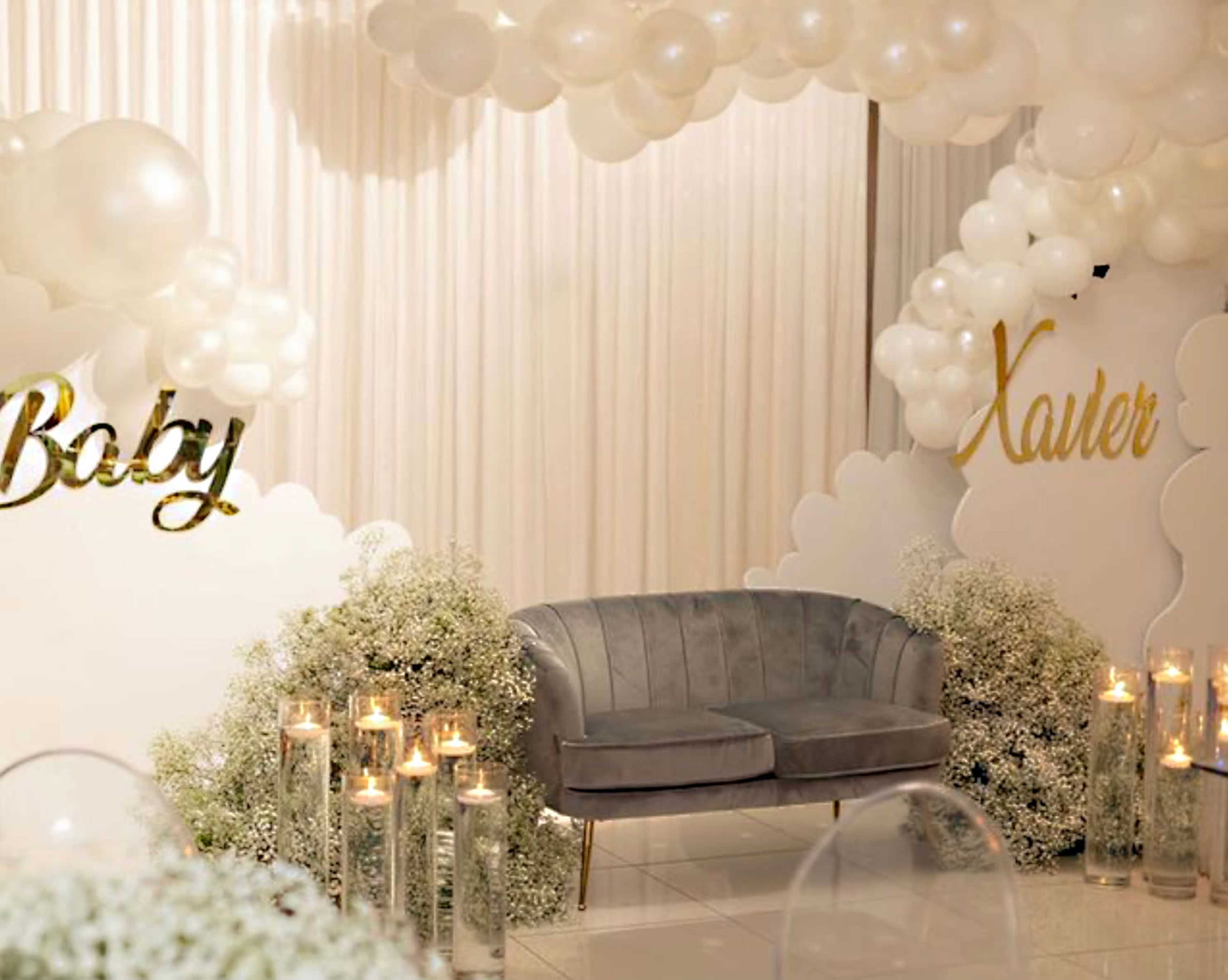 The image shows a decorated event space featuring a gray sofa surrounded by white balloons, floral arrangements, and lit candles.