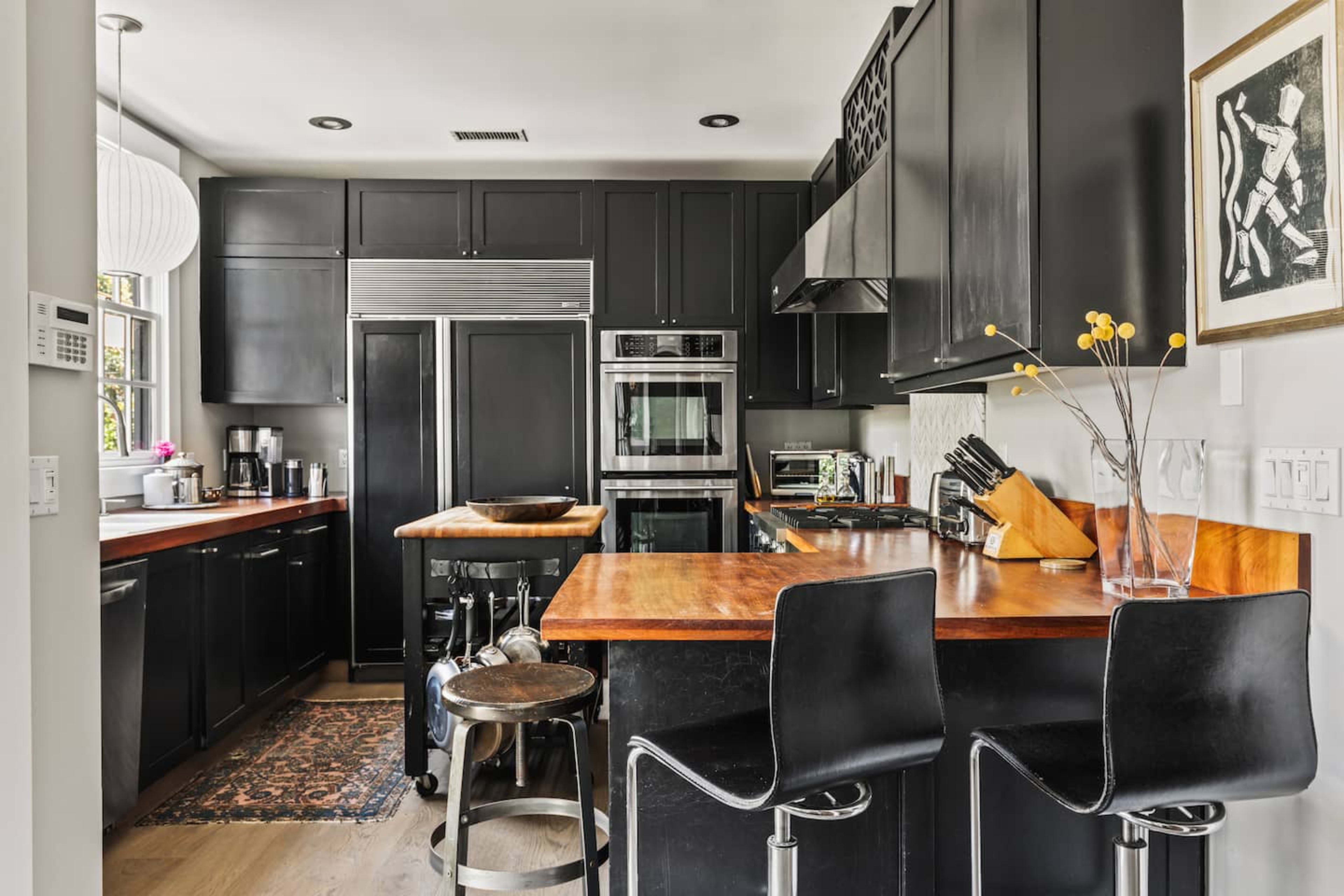 The image depicts a modern kitchen featuring black cabinetry, stainless steel appliances, and a wooden countertop with bar stools.