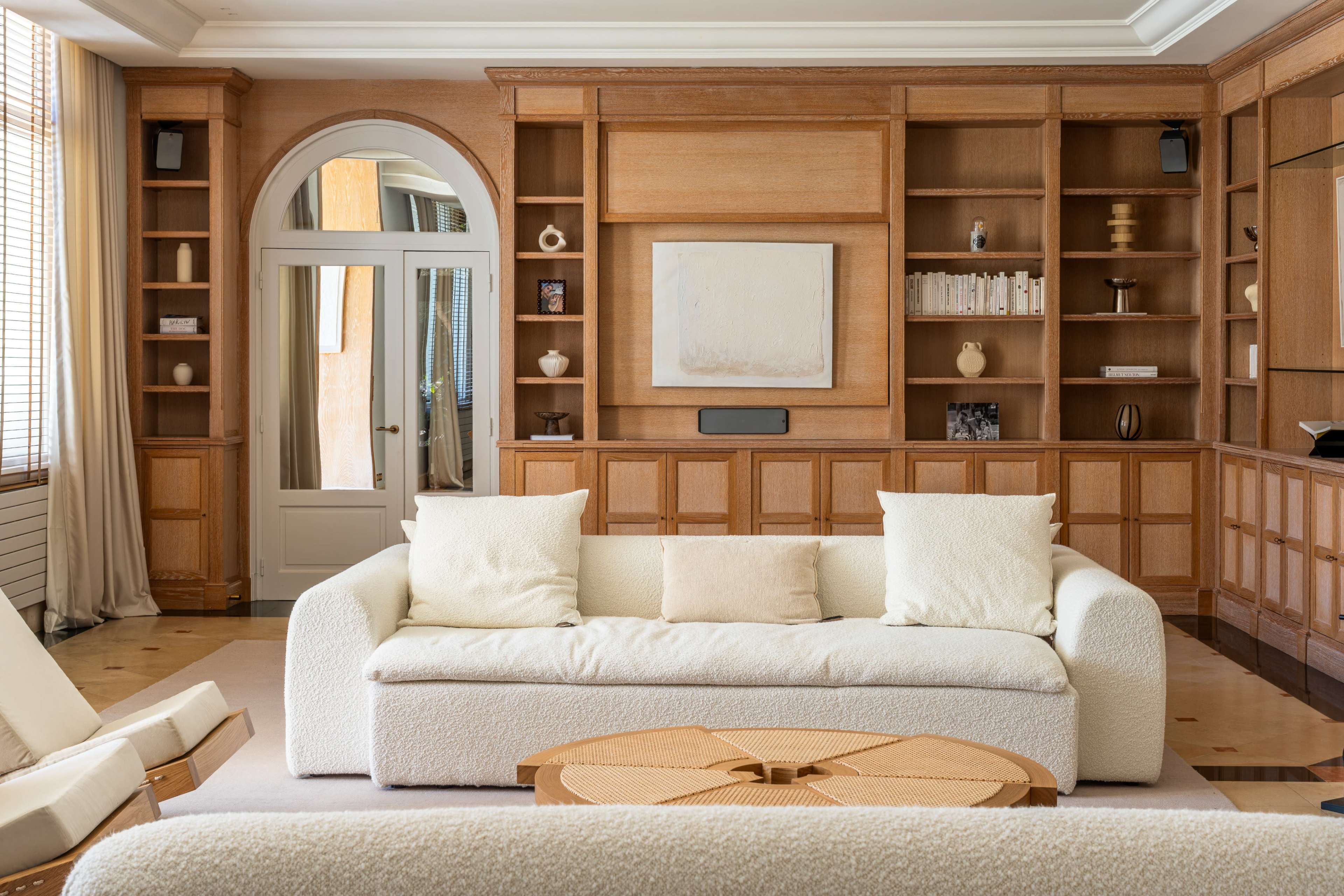 A spacious living room features a light-colored, textured sofa, a round coffee table, and wooden built-in shelves filled with decorative items.