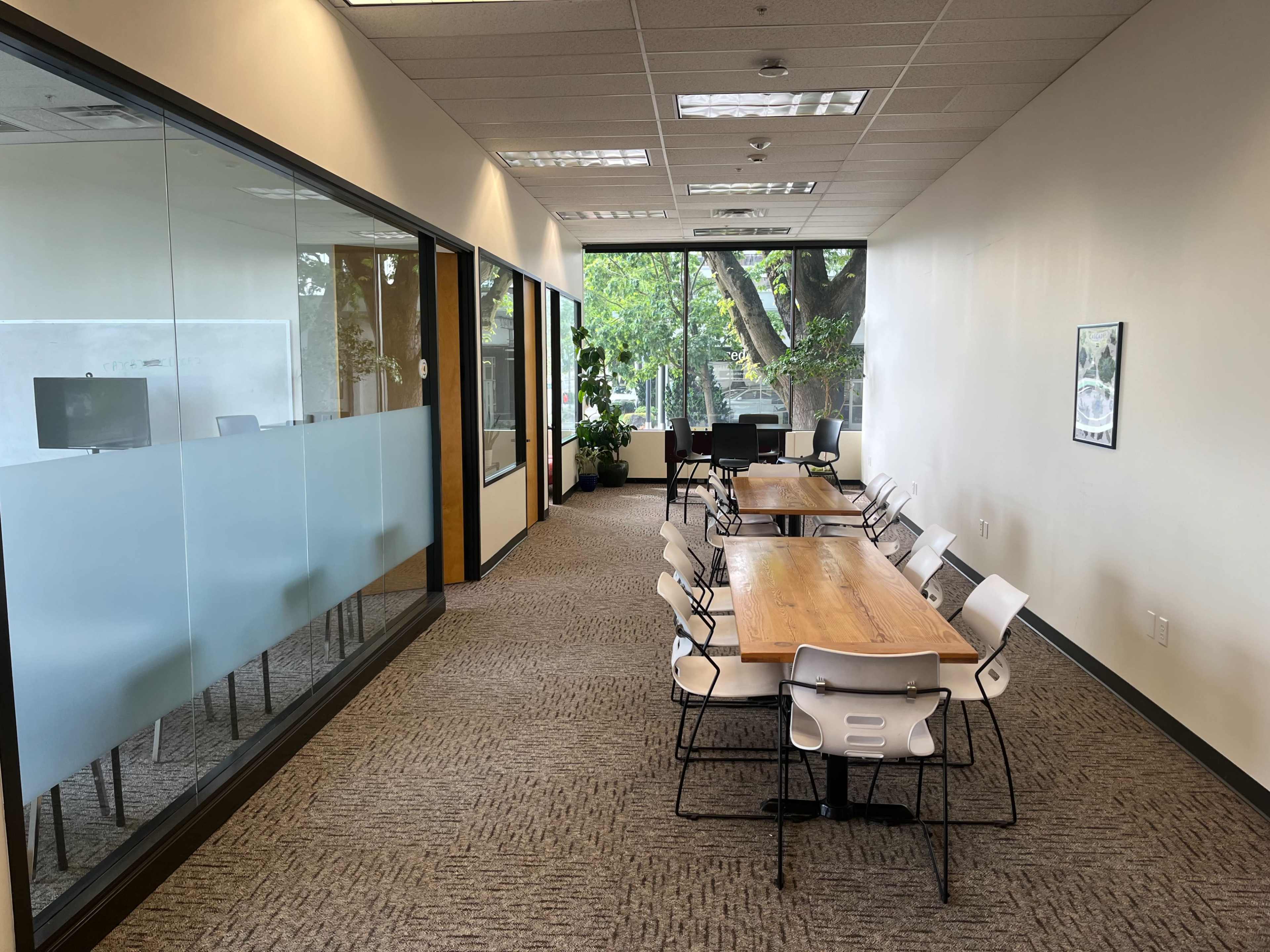 The image shows a hallway in an office environment featuring a set of wooden tables and chairs, along with glass-walled meeting rooms and greenery visible through the windows.