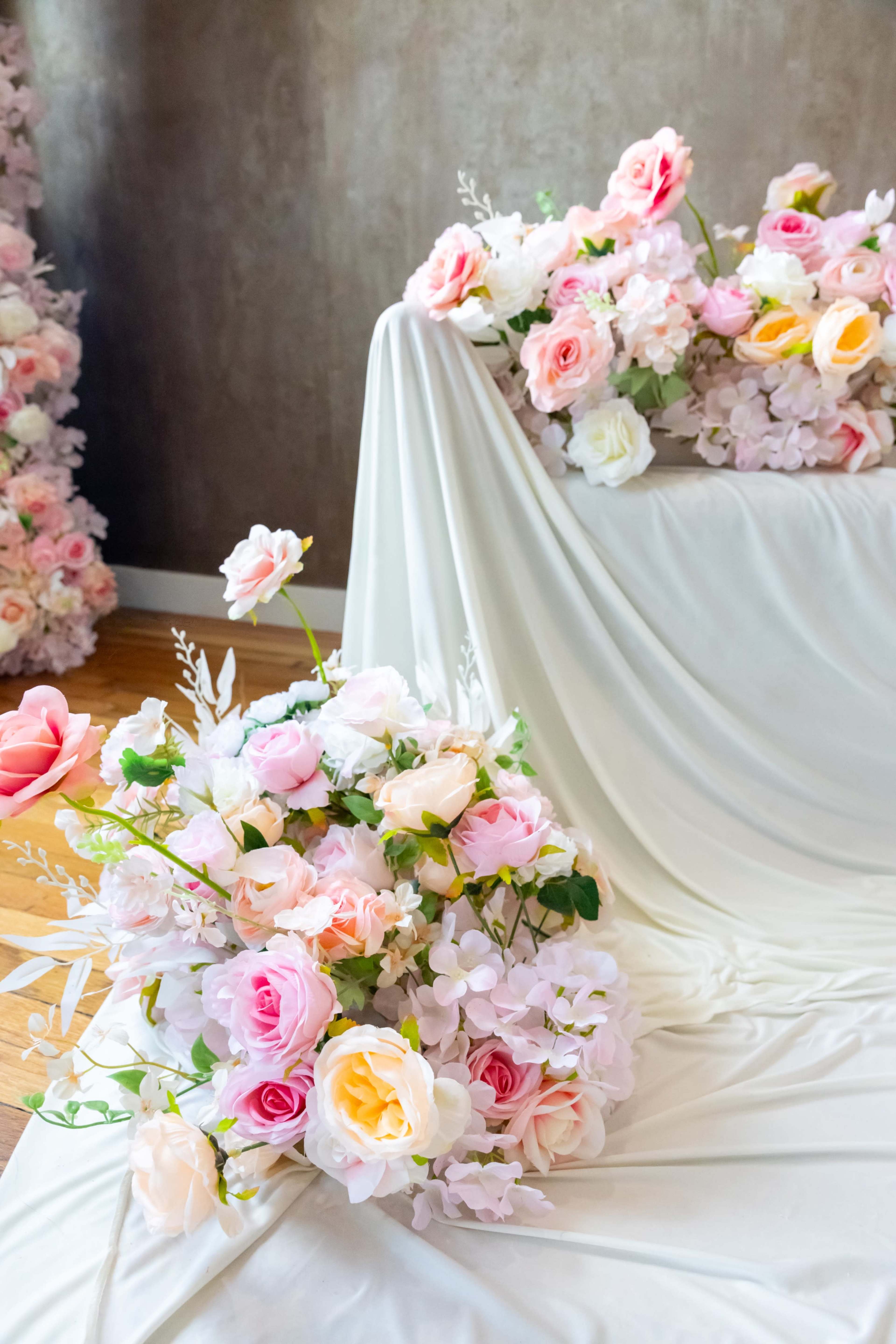 A variety of artificial flowers in soft pinks and whites are arranged on a white draped surface, with additional flower arrangements placed in the background.