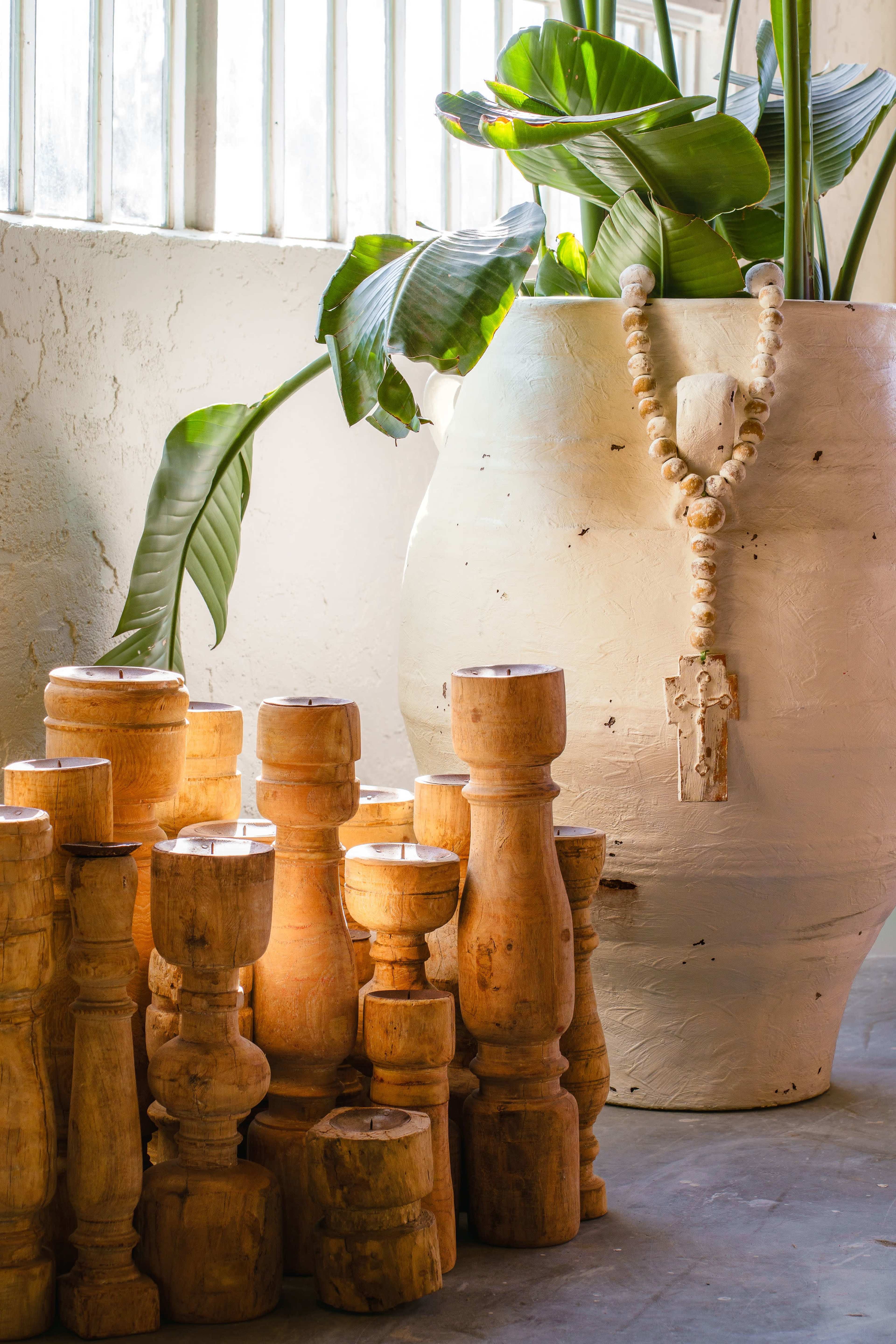 The image features a collection of wooden cylindrical objects stacked next to a large white ceramic pot adorned with greenery and a beaded necklace.