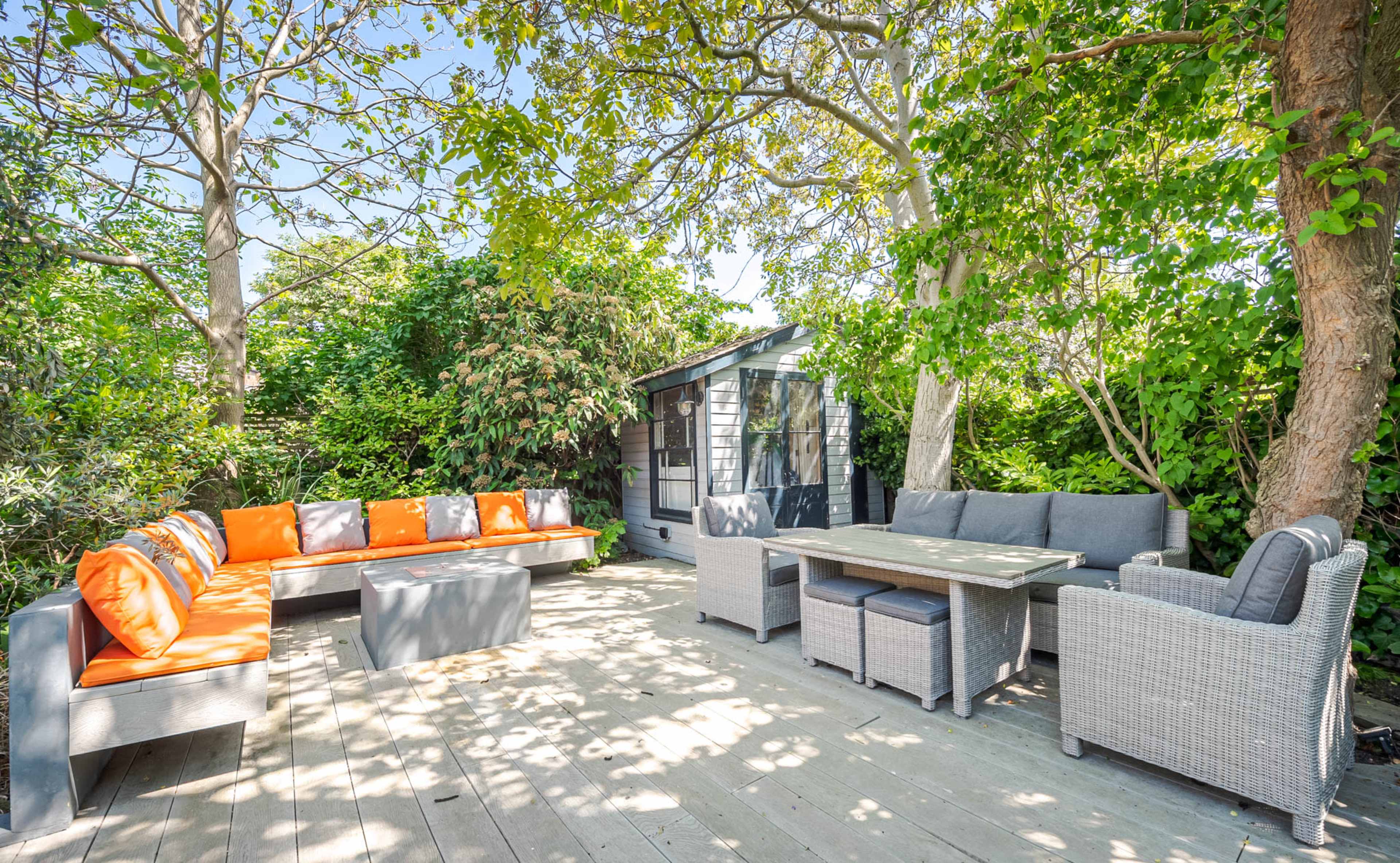 A patio area features a seating arrangement with orange and gray cushions, surrounded by lush greenery and a small shed in the background.