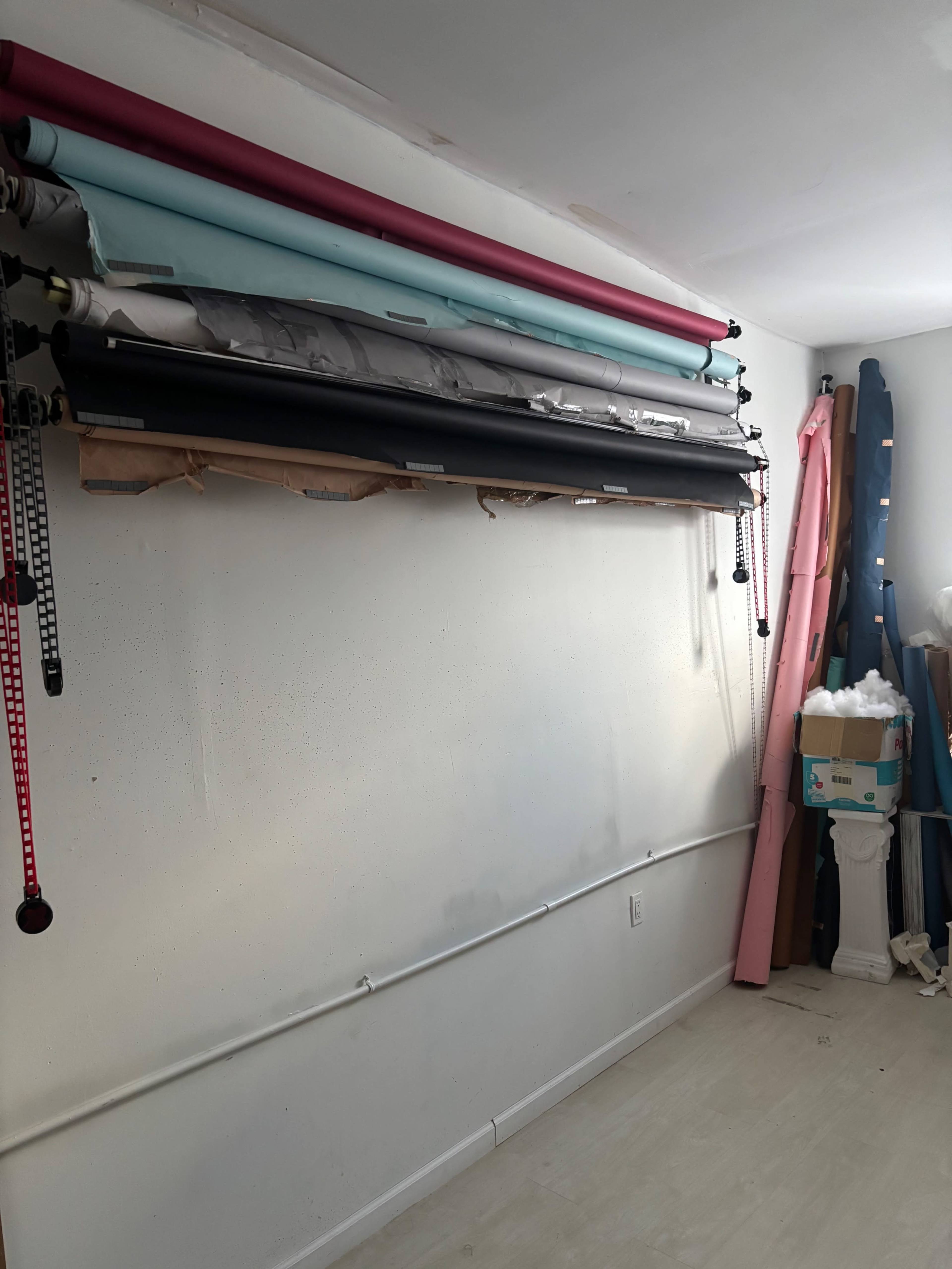 The image shows a wall with several rolls of colored backdrop paper hanging above a light-colored floor, alongside a stack of materials and equipment in the corner.