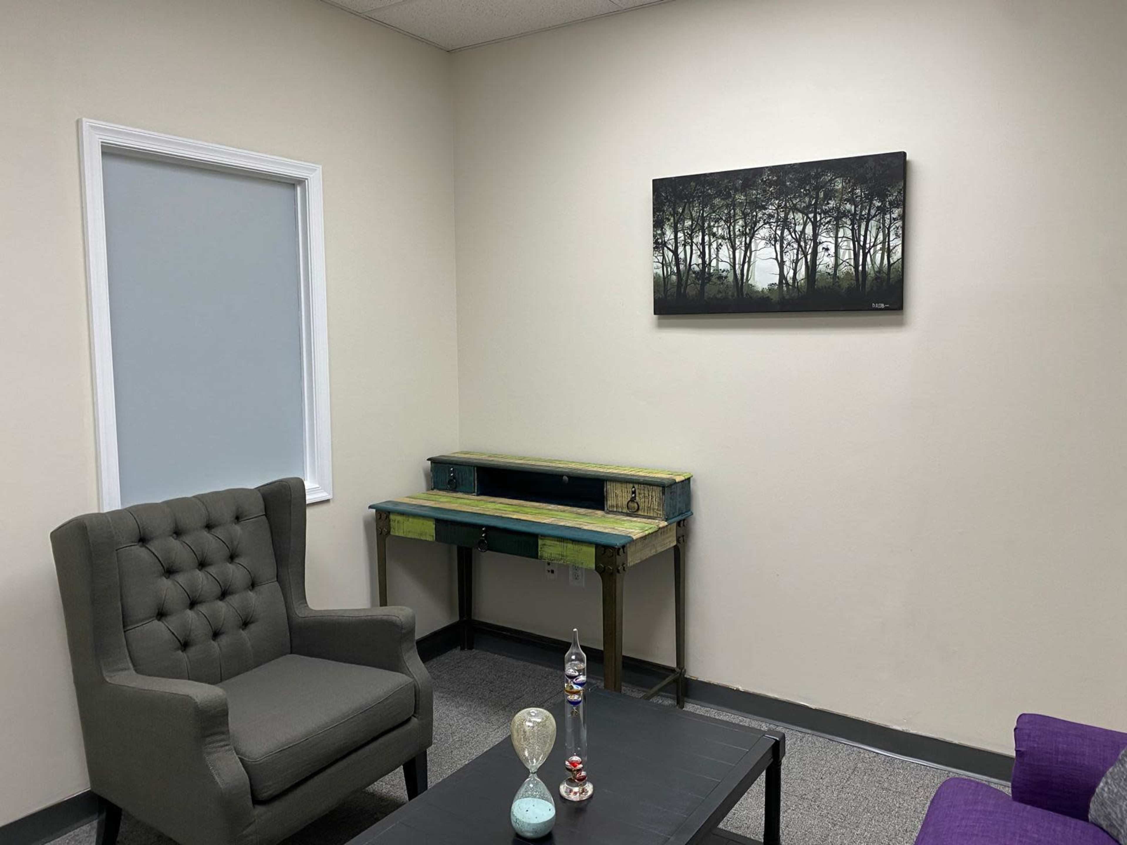 The image shows a simple room with a gray armchair, a wooden desk against the wall, and a painting of trees hanging above it.