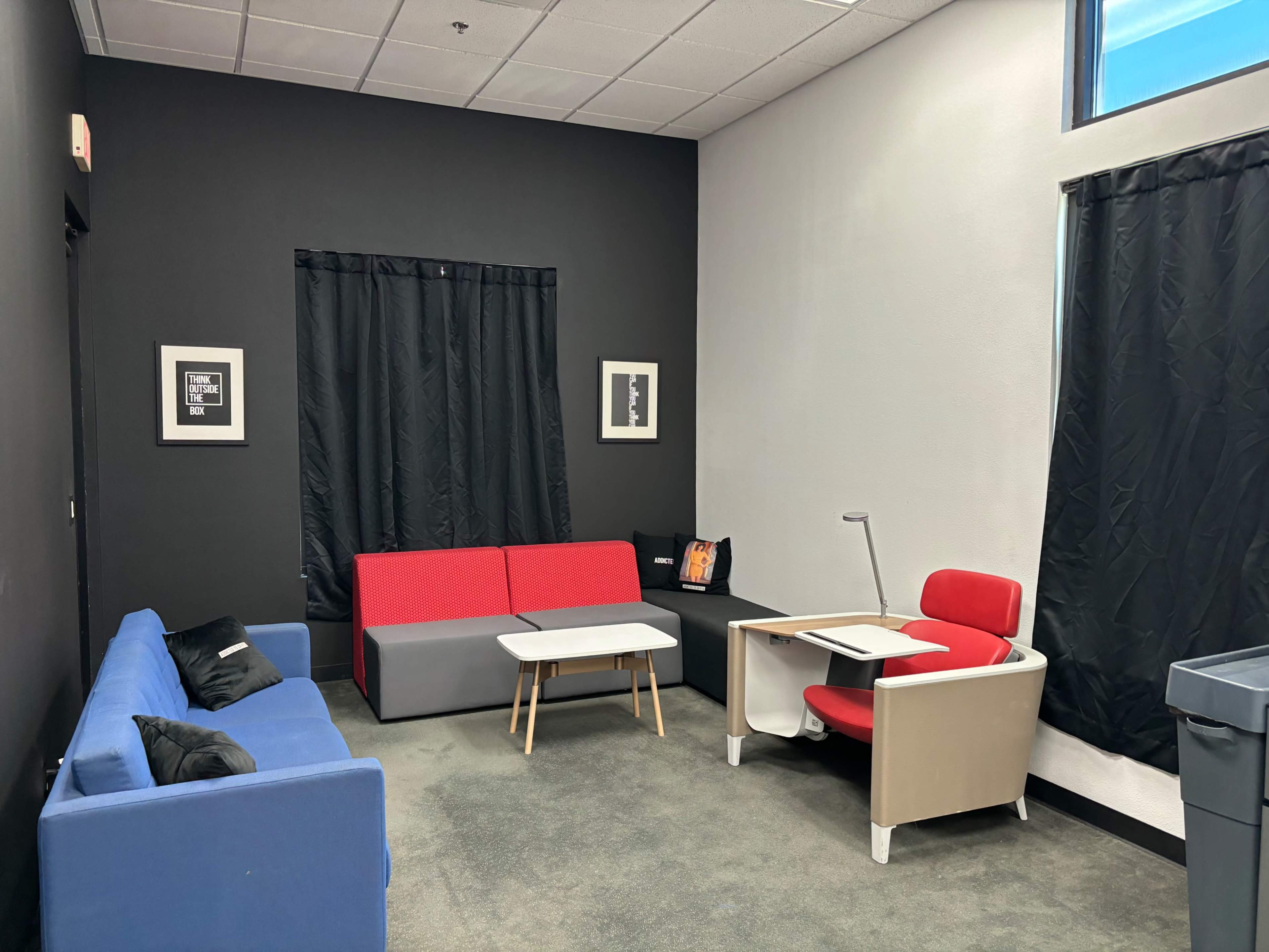 The image shows a small lounge area featuring two sofas, a coffee table, and a desk with a chair, all set against a dark wall with a window covered by a black curtain.