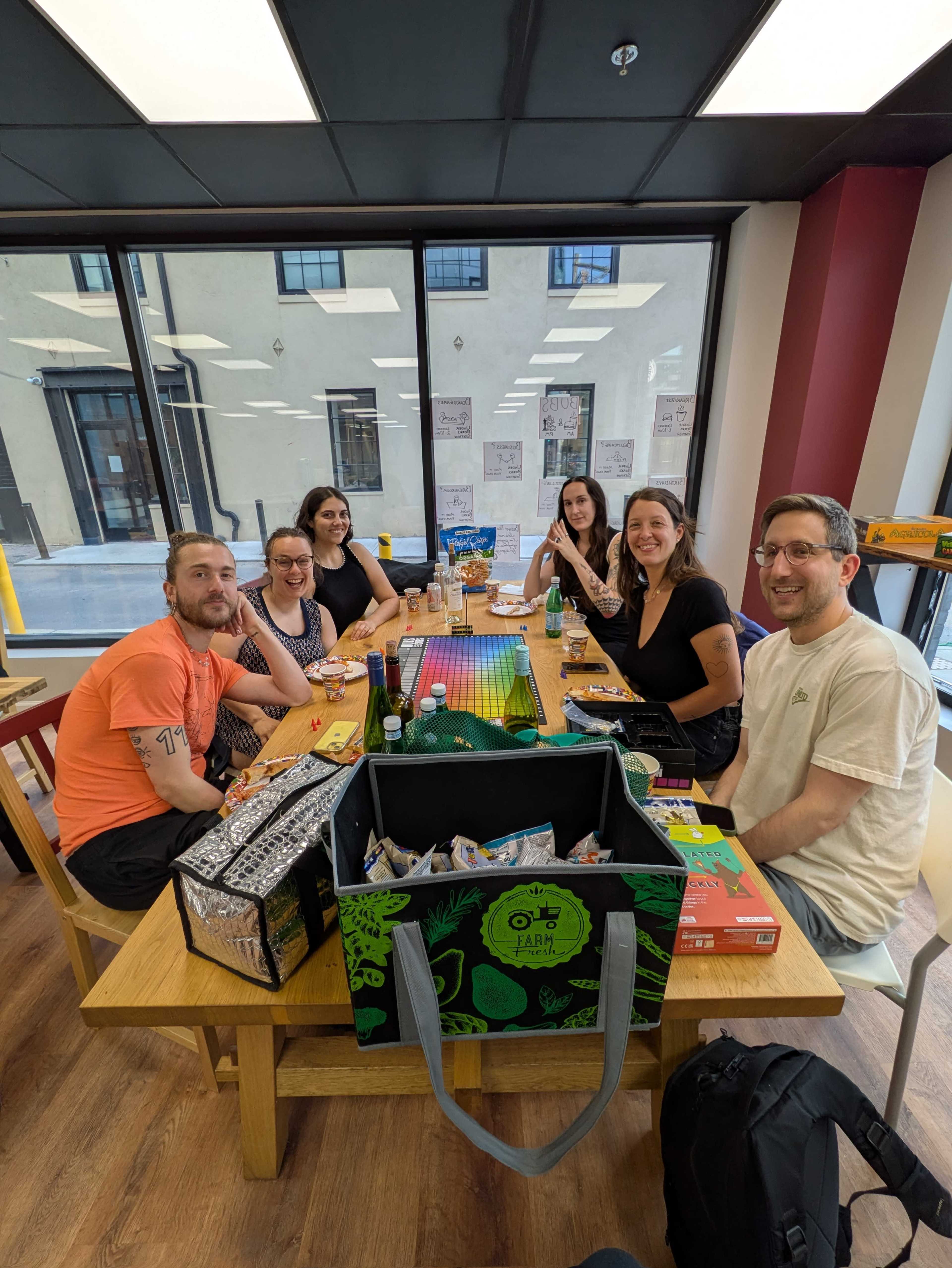 A group of six people sits around a table in a bright room, enjoying snacks and playing games.