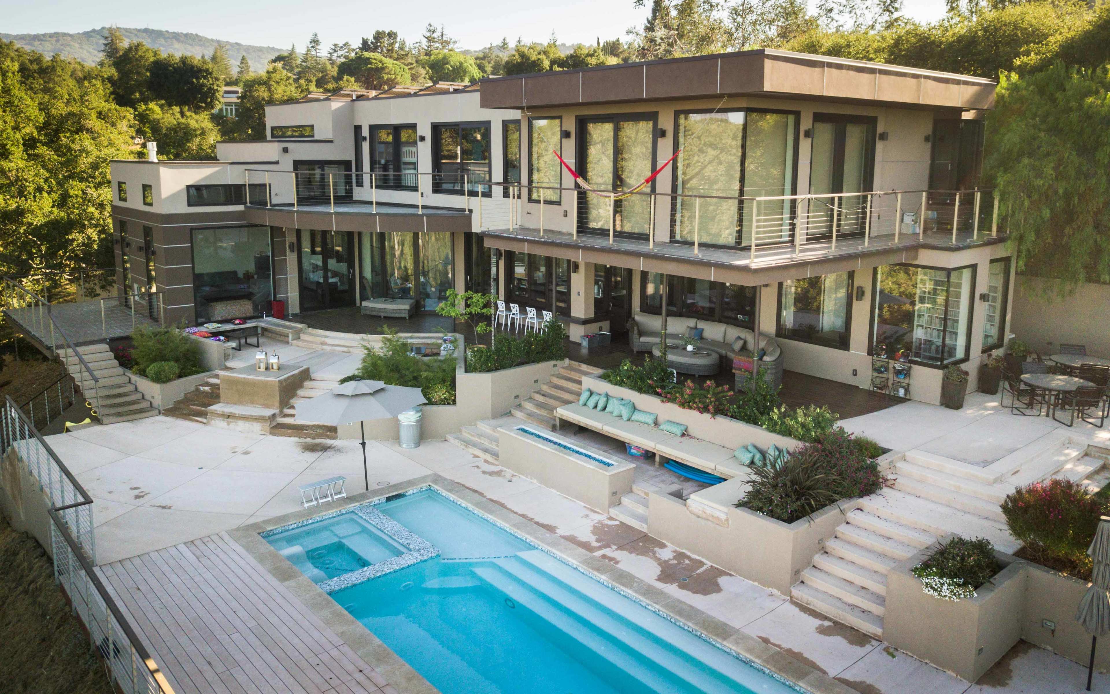 A modern house with a pool and multiple outdoor seating areas is surrounded by greenery.