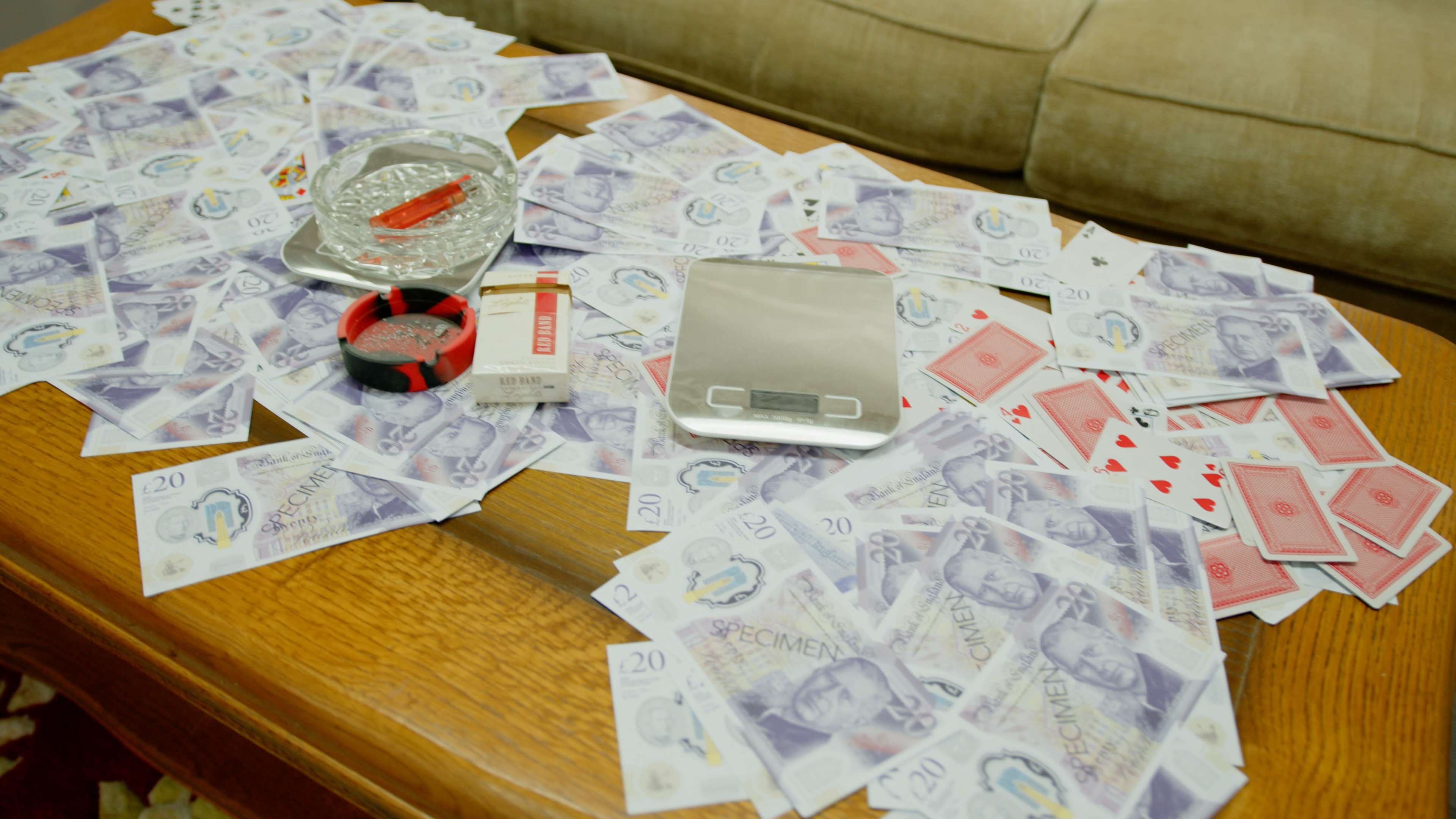 The image shows a wooden table covered with large amounts of banknotes, a scale, playing cards, and other items.