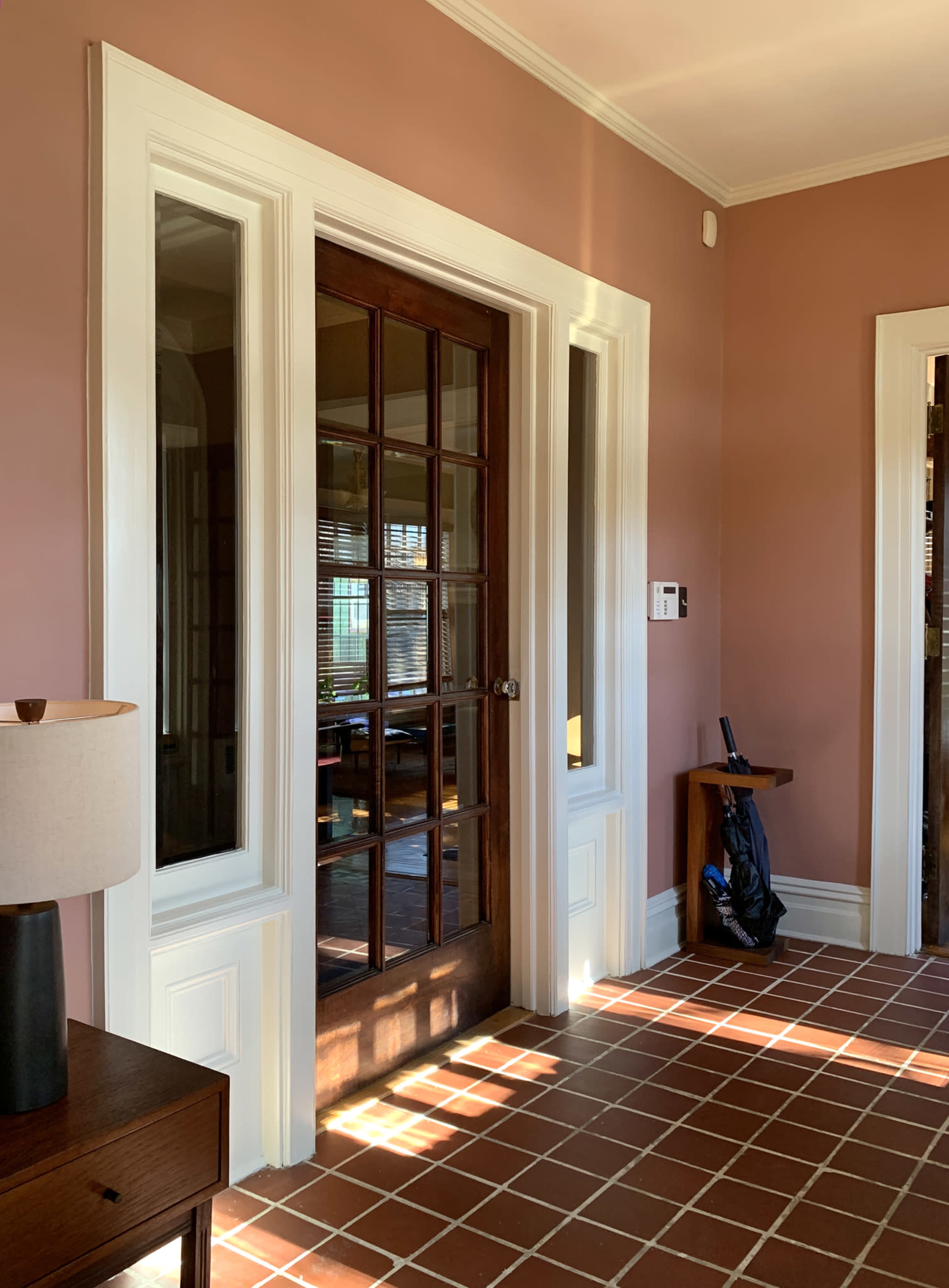 A hallway with a wooden door featuring glass panels, flanked by windows, and a tiled floor illuminated by sunlight.