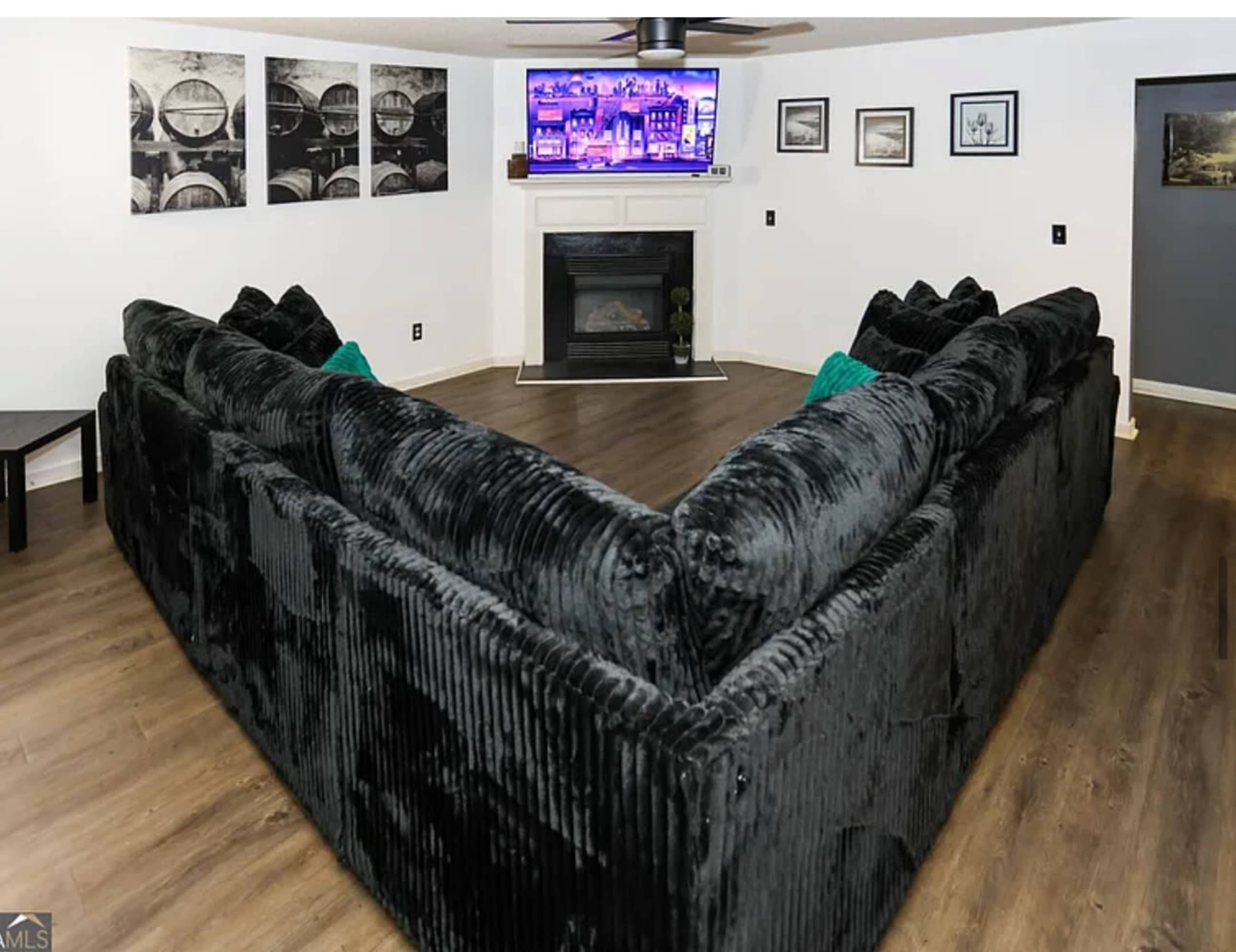 A large black sectional sofa is positioned in an L-shape around a wall-mounted TV and a fireplace in a living room with light-colored walls and wooden flooring.