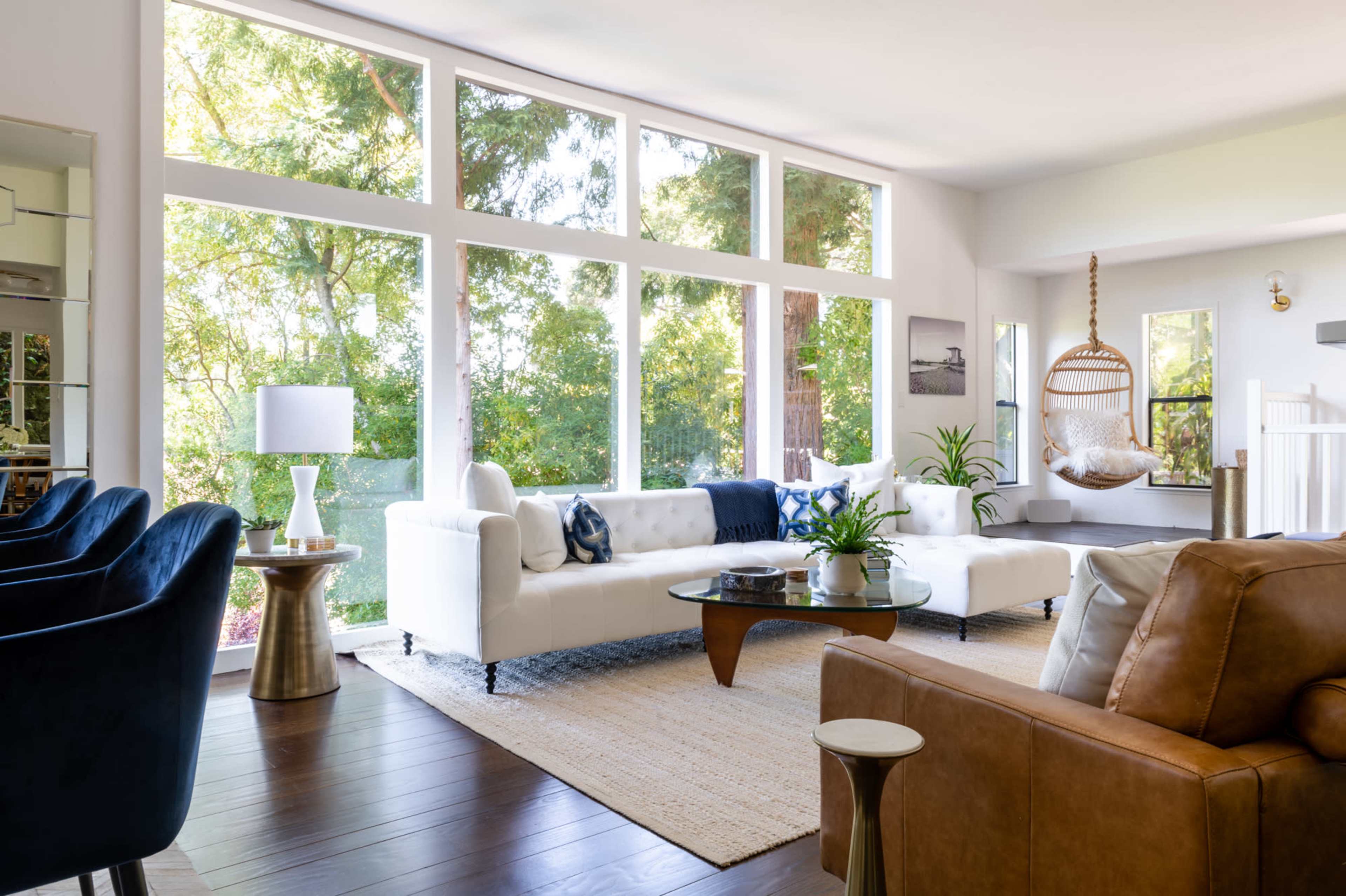 The image shows a bright living room with large windows showcasing greenery outside, featuring a white sofa, a glass coffee table, and a hanging chair.
