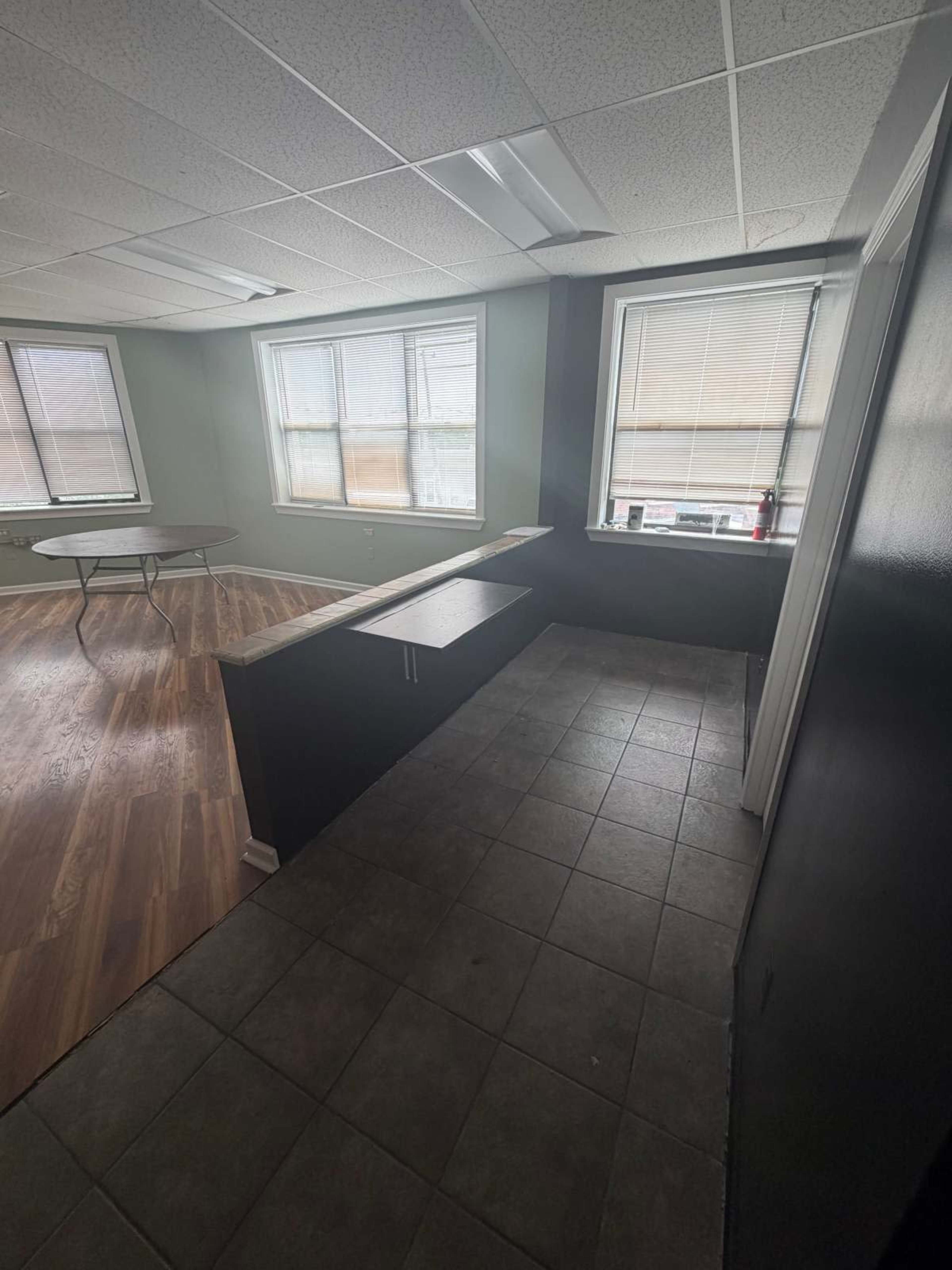 The image shows a room with large windows, a tiled floor, and a countertop area beside a dark wall, with a table in the background.