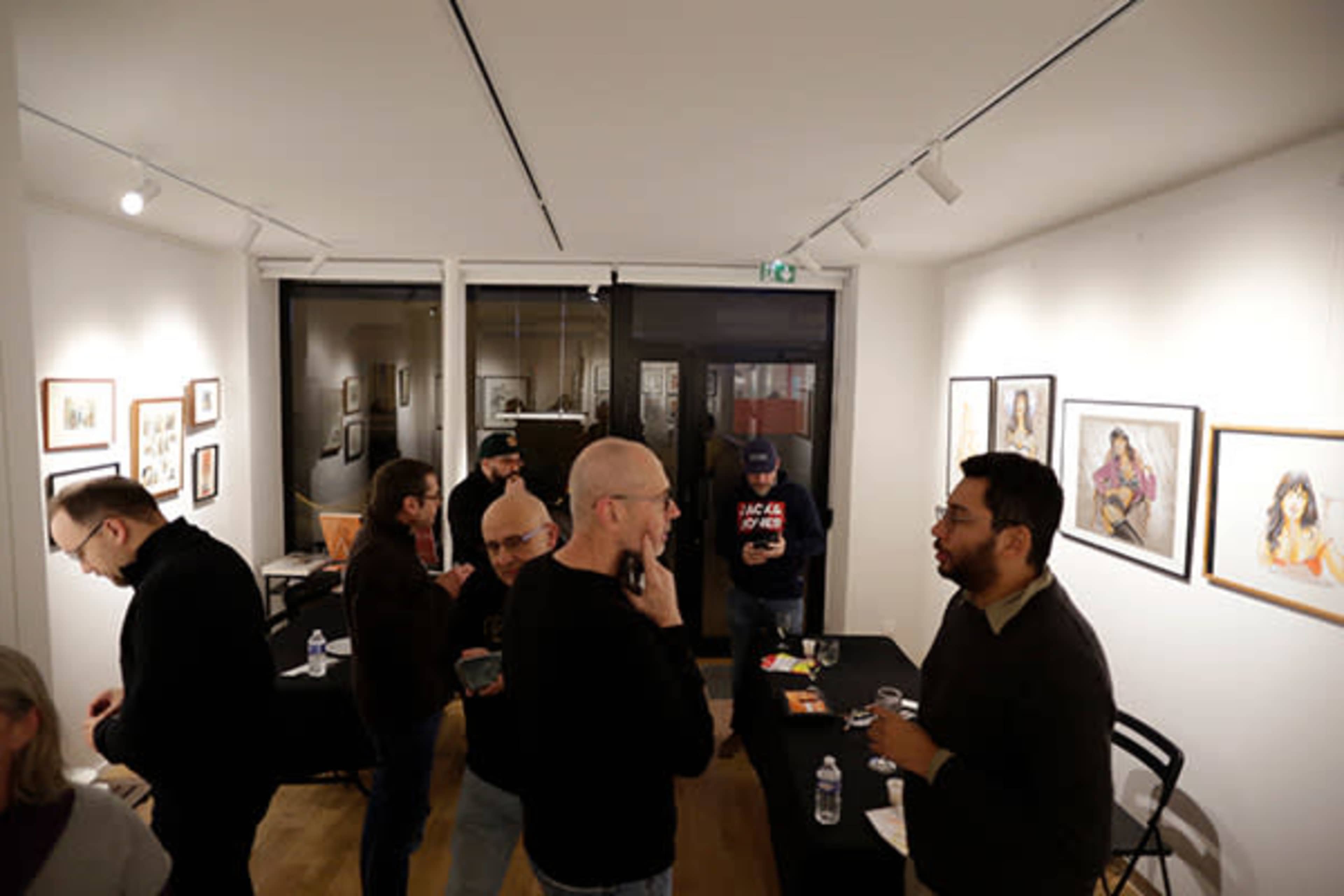 A group of people is gathered in an art gallery, engaging in conversation amidst framed artwork on the walls.