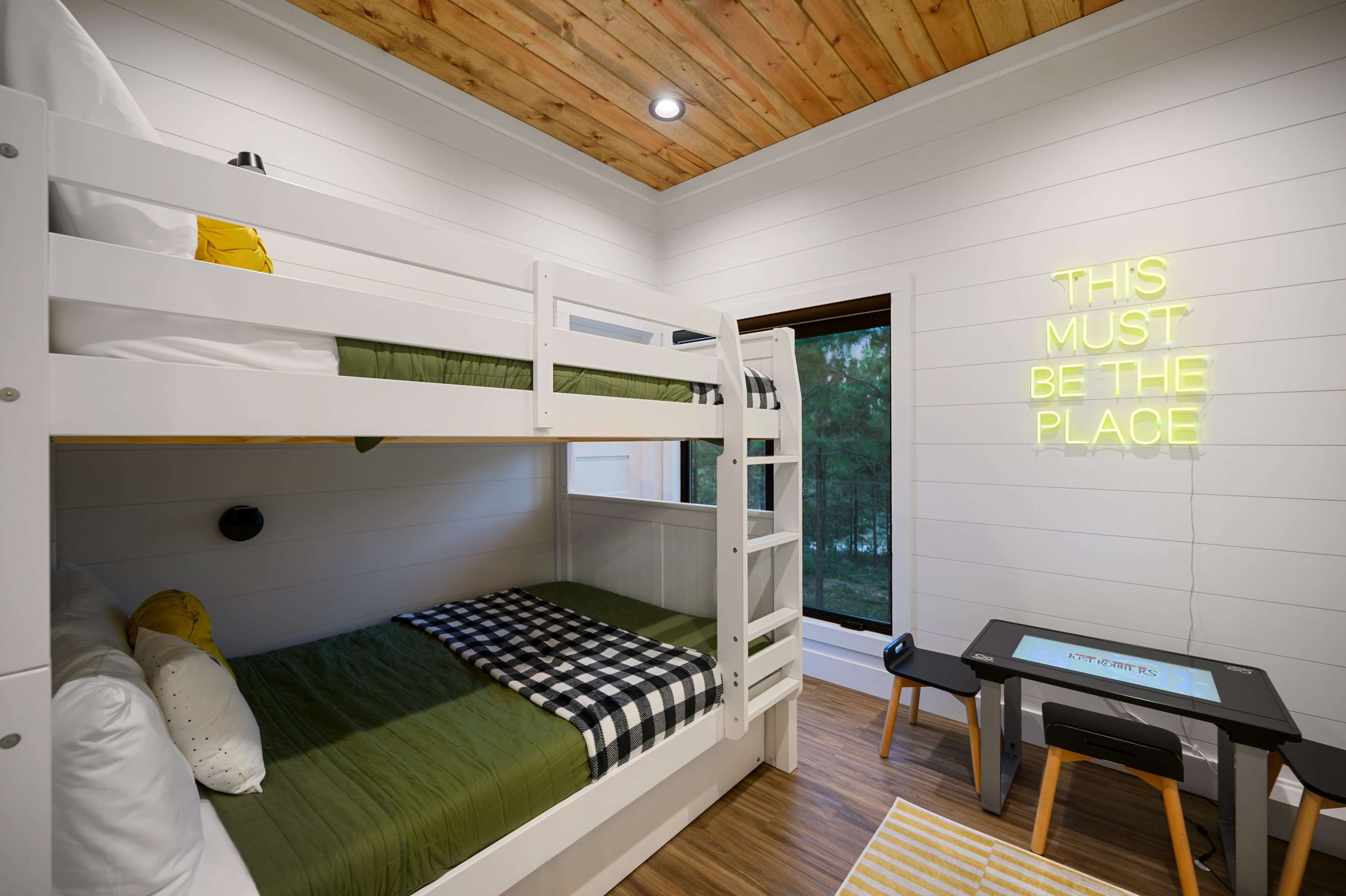 A bedroom featuring a bunk bed with green and black bedding, a small desk, and a neon sign on the wall.