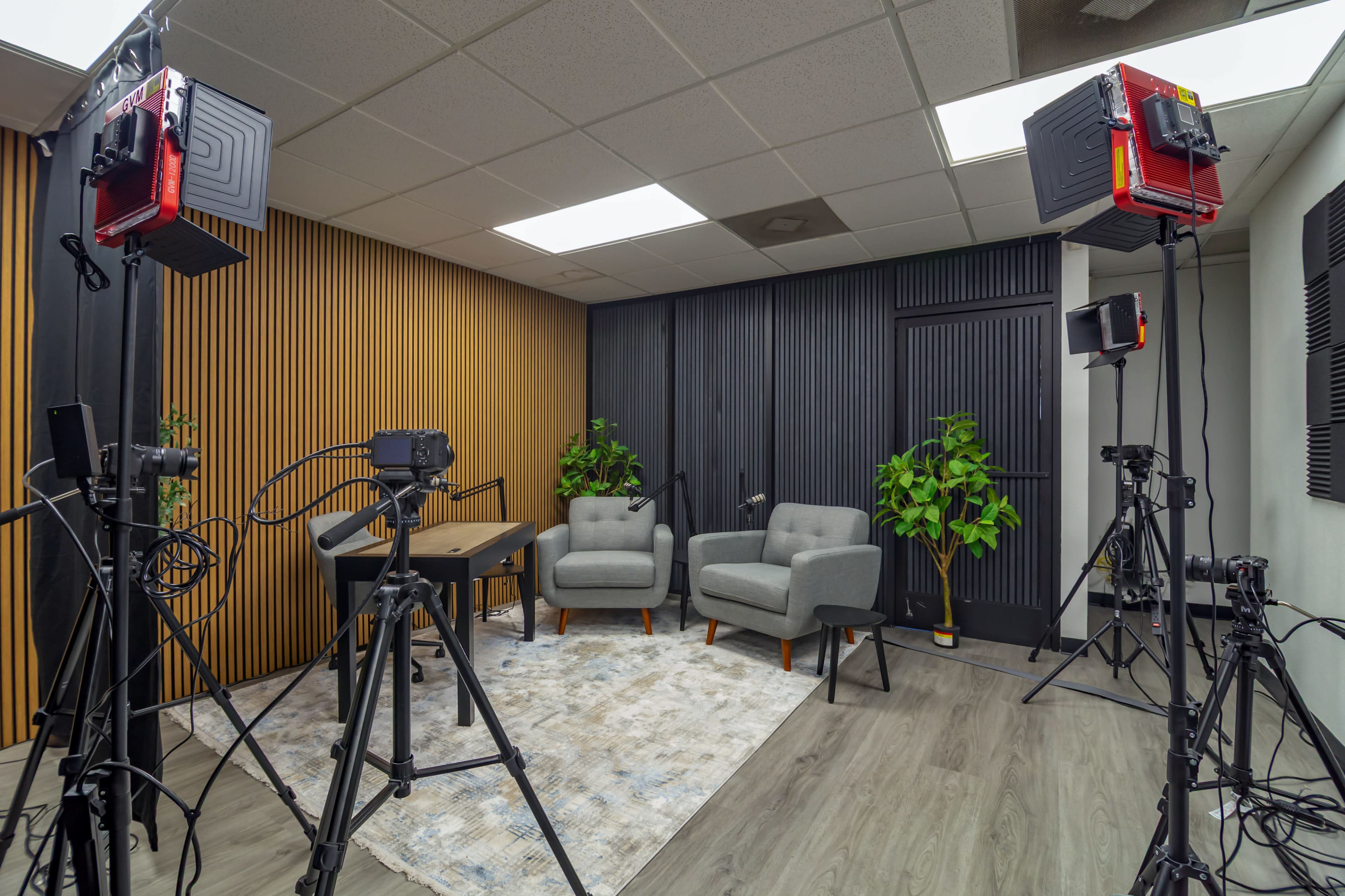 The image shows a modern studio setup with two gray armchairs, a wooden table, and several camera tripods and lighting equipment arranged around it.