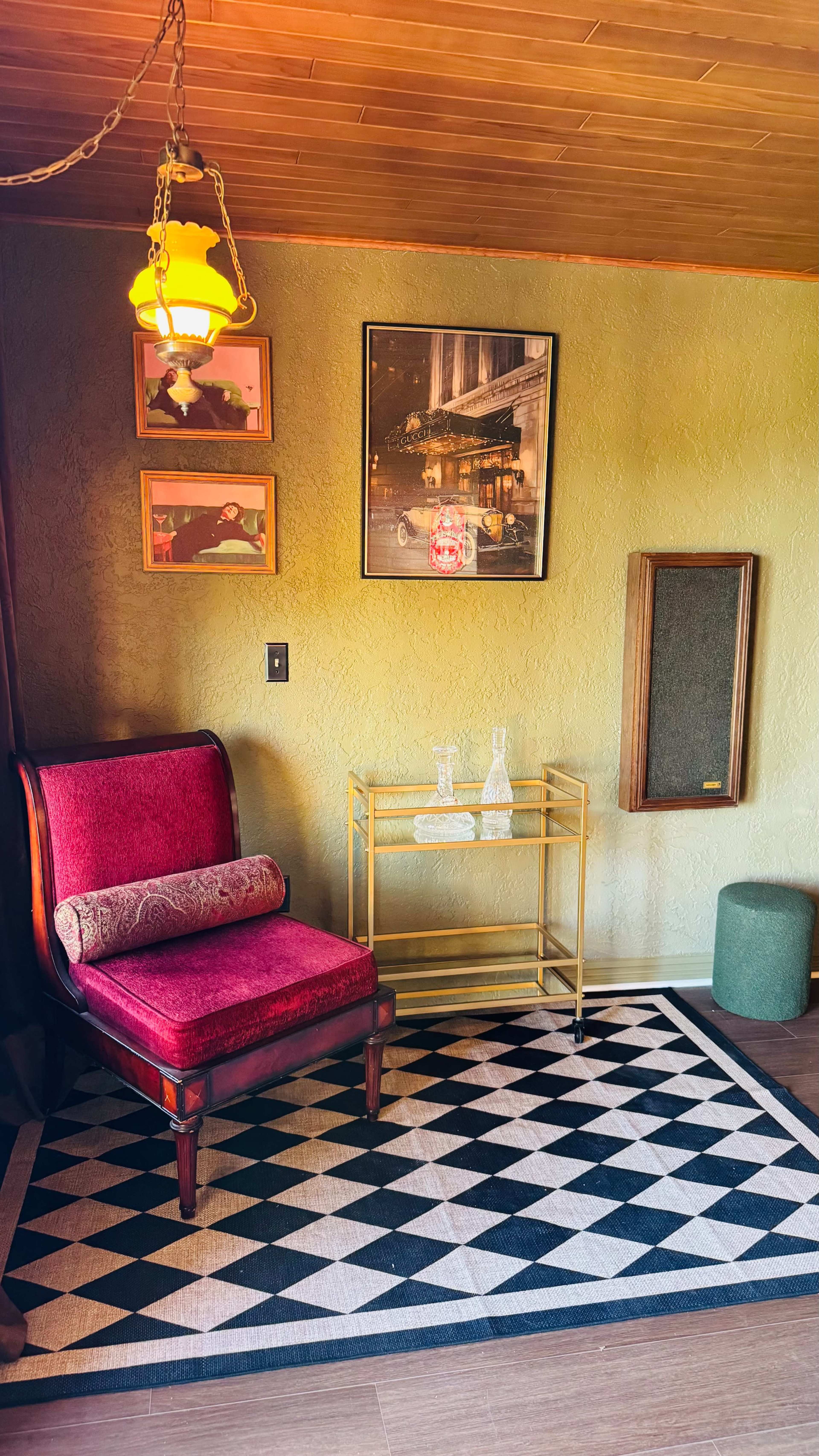 The image shows a corner of a room featuring a red upholstered chair, a gold serving cart with glassware, and a patterned rug on the floor, along with wall decorations including framed photos and a light fixture.