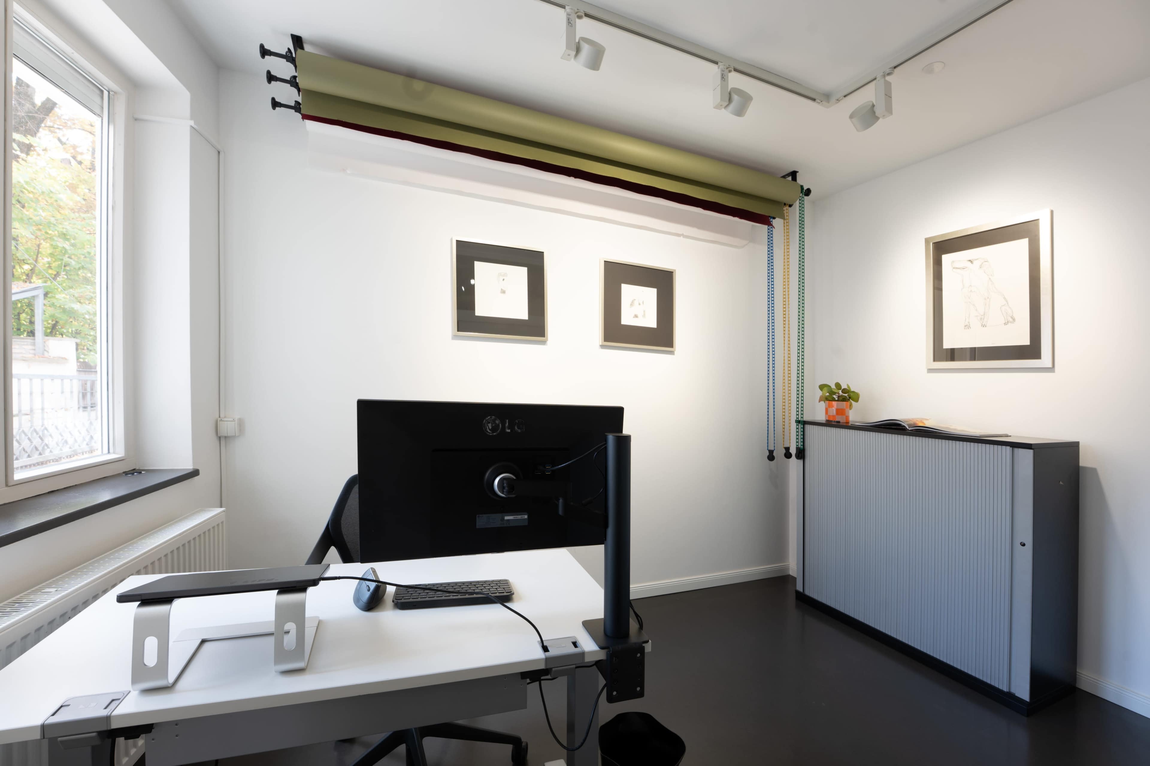 A modern office space features a desk with a computer, artwork on the walls, and a storage cabinet.