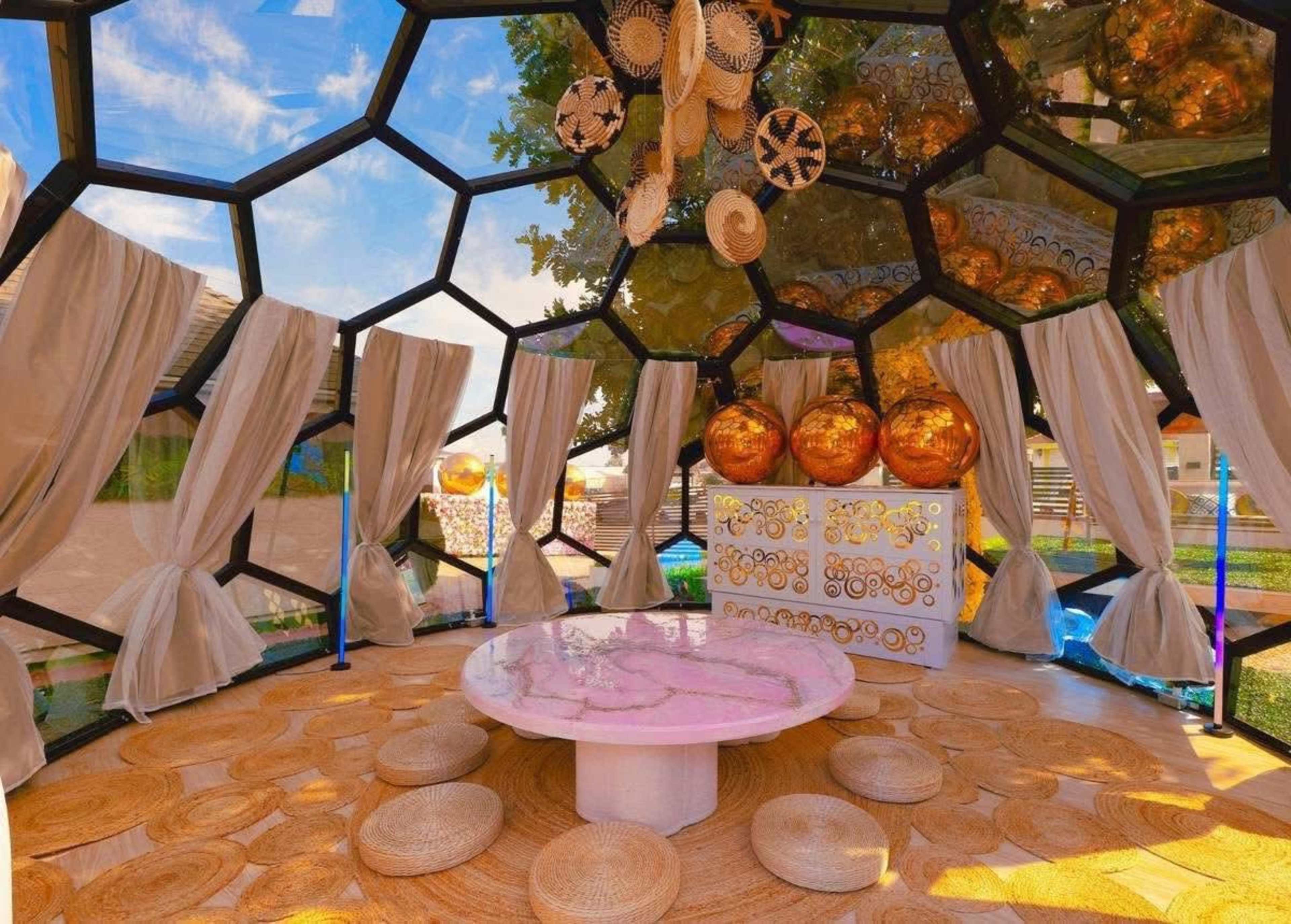 The image shows a modern, hexagonal pavilion with a round pink table, surrounded by round seating and decorative golden ornaments, illuminated by natural light.